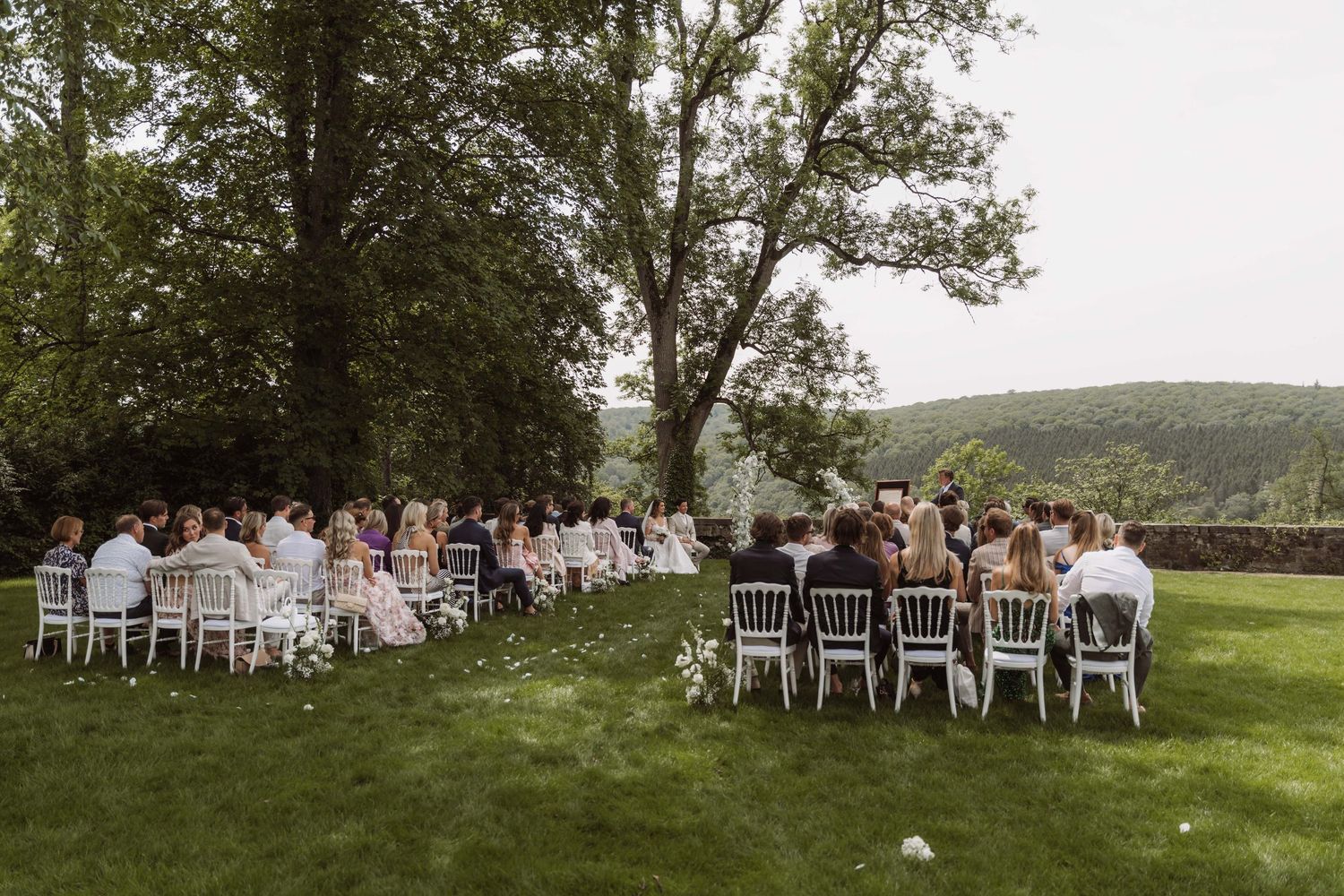 Wedding guests seated in white chairs on a grassy hillside overlooking a scenic mountain landscape during an outdoor ceremony.