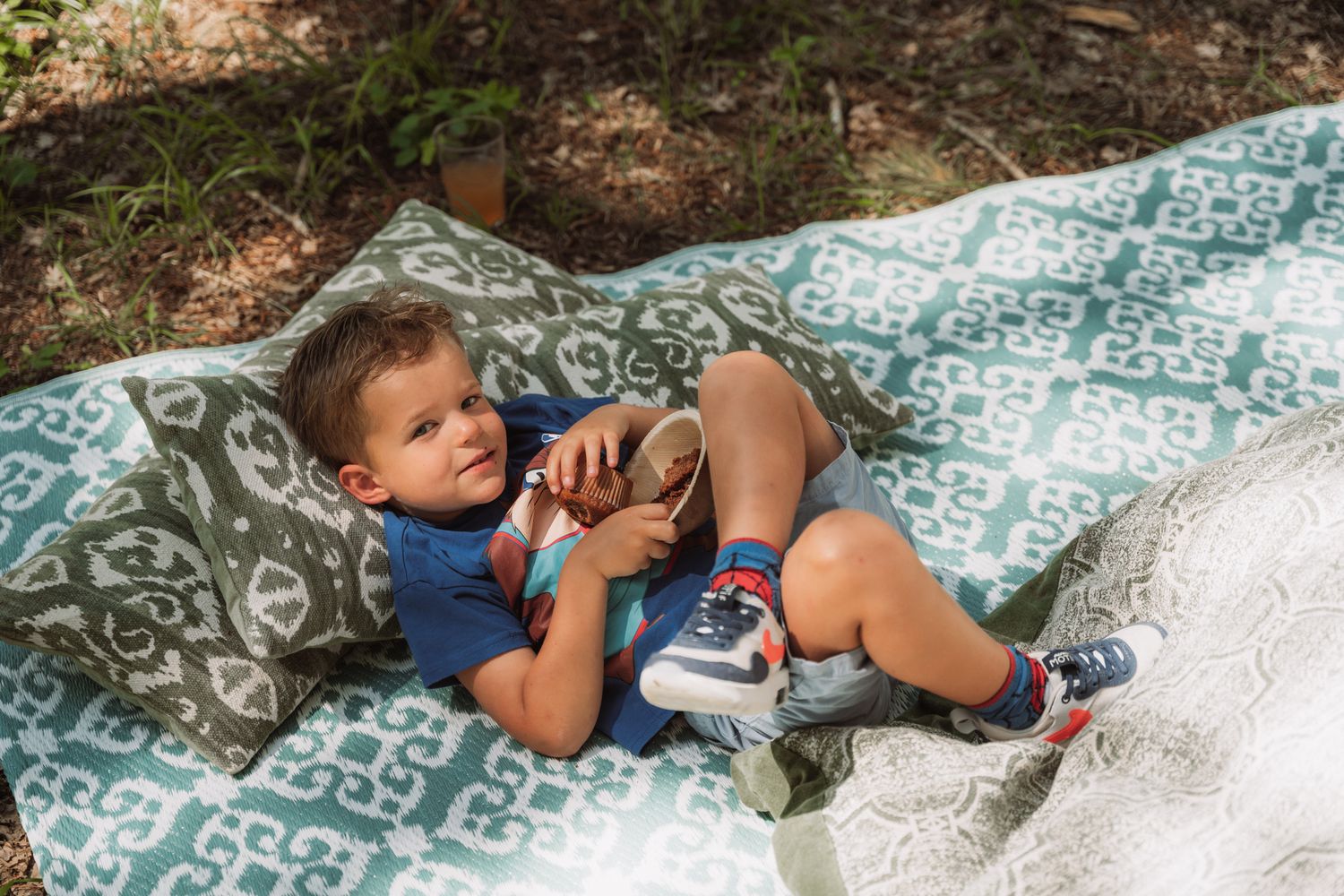 A young child relaxes on a patterned blanket outdoors while holding a mobile device on a sunny day.