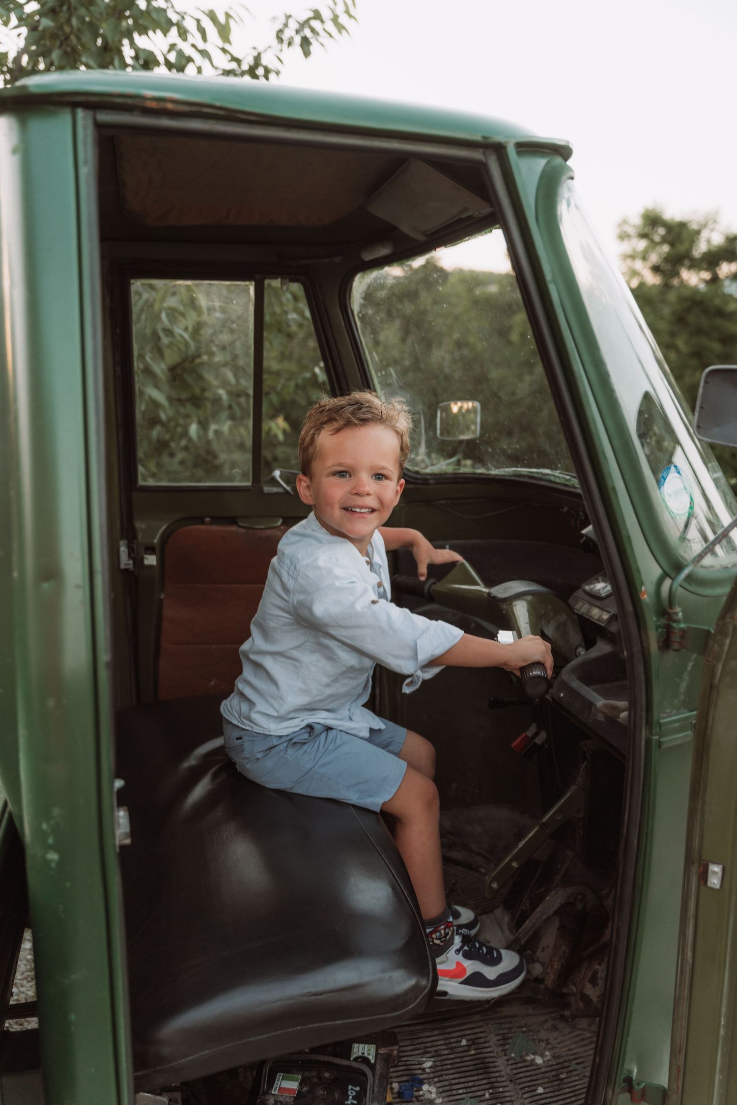 A young child sits in the driver's seat of a vintage green truck, smiling while pretending to drive.