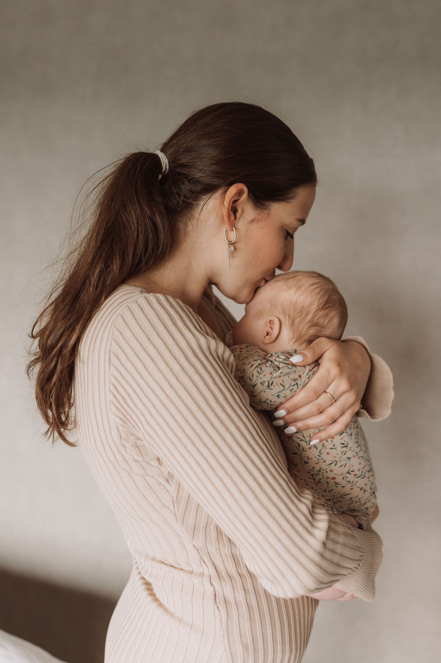 Mother in beige sweater holding and kissing baby in soft natural light.