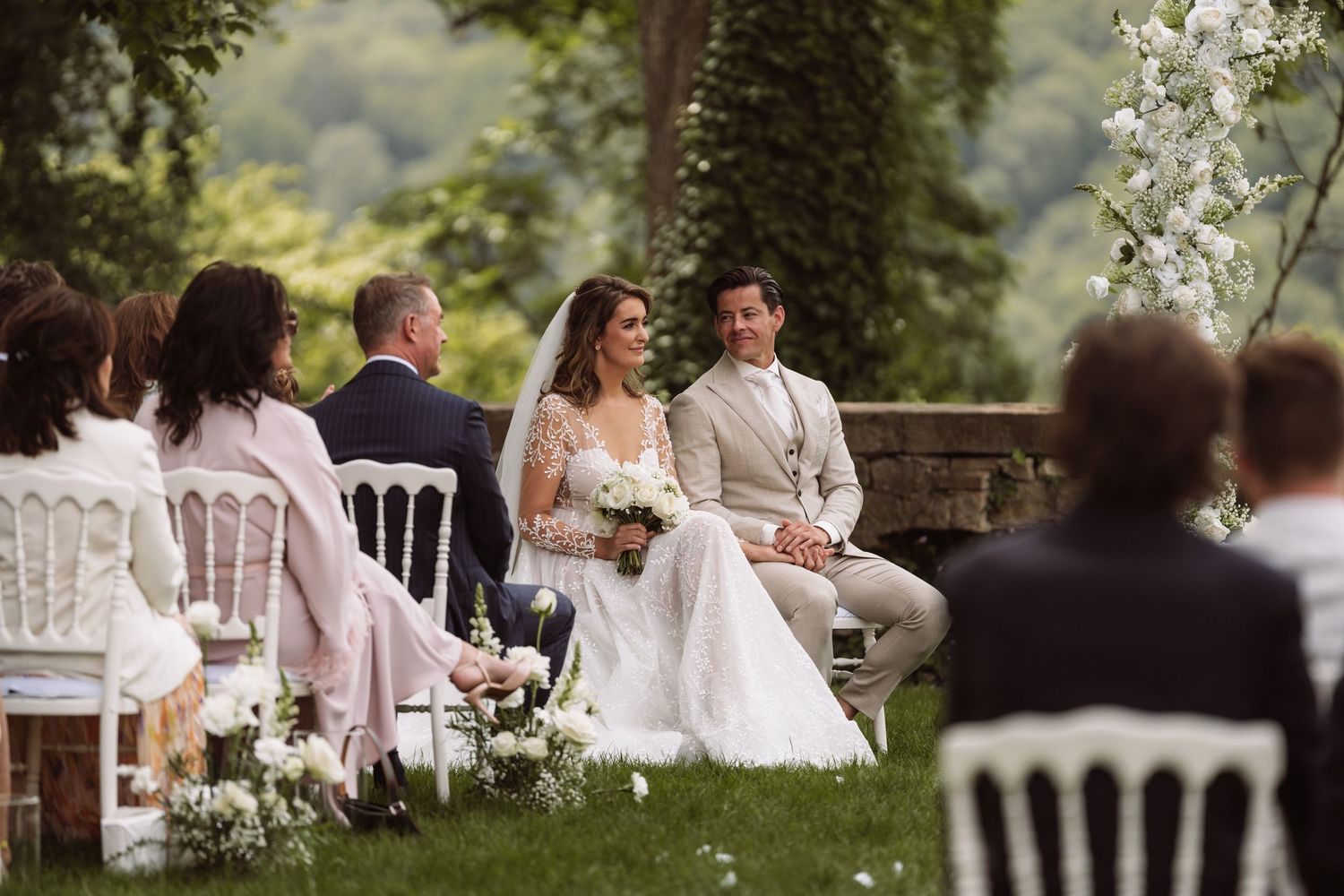An outdoor wedding ceremony with white chairs and floral decorations in a garden setting.