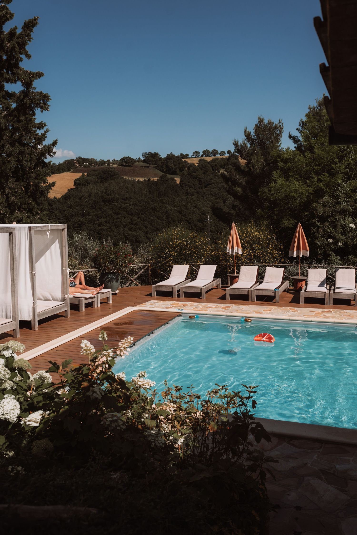 Luxury outdoor swimming pool with white lounge chairs and orange umbrellas surrounded by lush landscaping and hills.