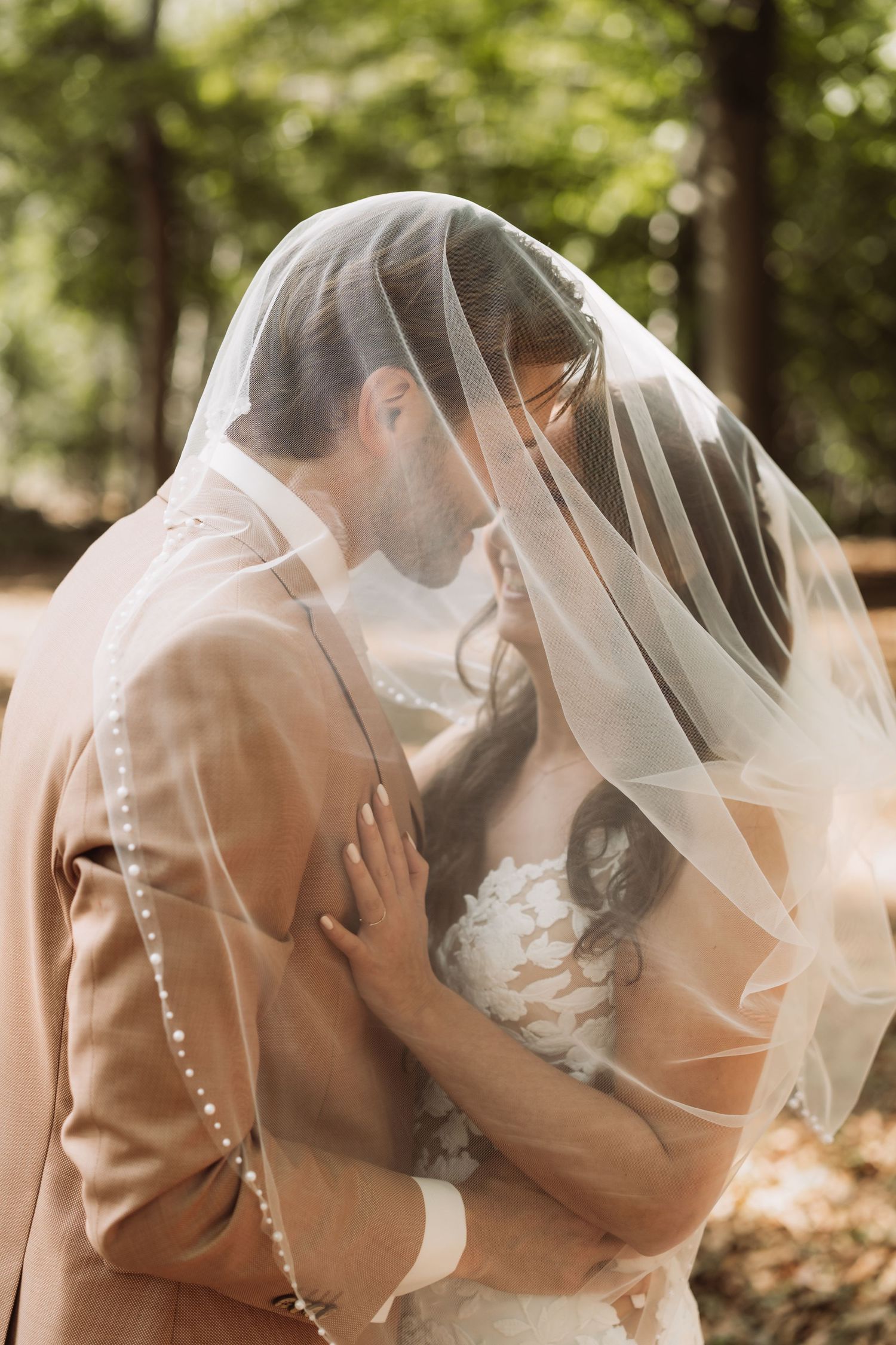 Romantic wedding couple shares tender kiss under flowing white veil in sunlit outdoor forest setting.