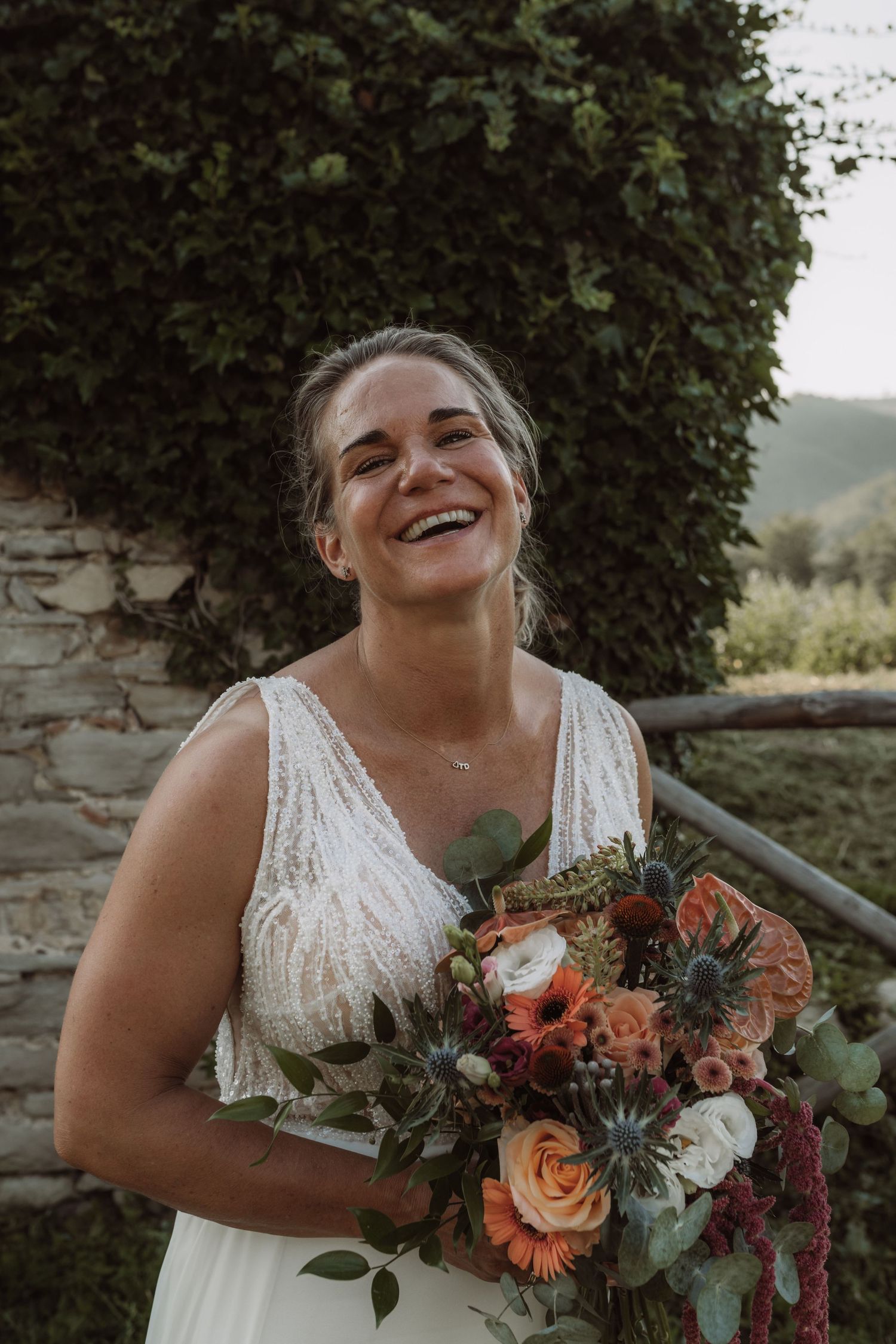 A joyful bride in a white lace dress holds a vibrant autumn bouquet with orange and burgundy flowers.