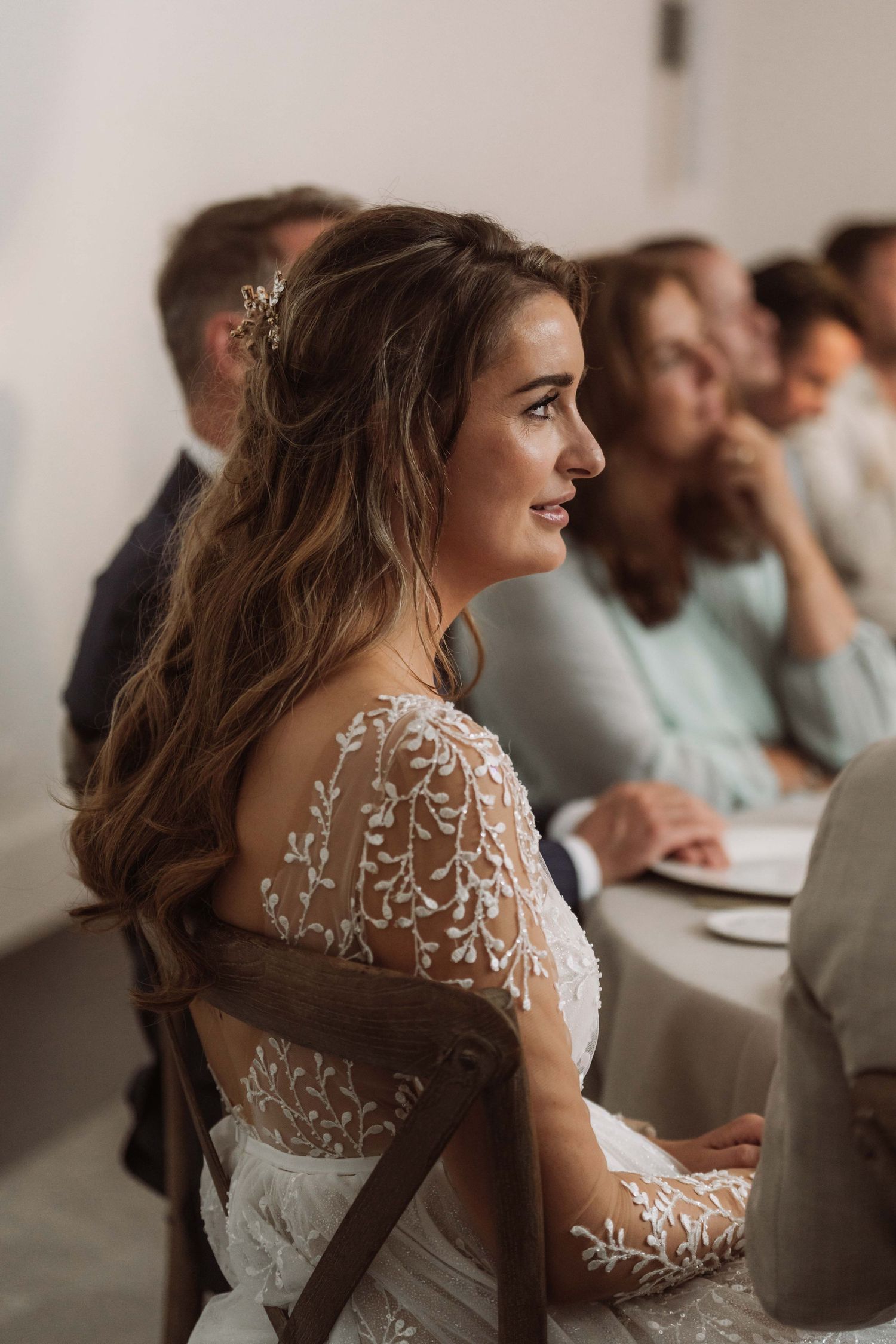 A bride in a white lace wedding dress with sheer sleeves sits among wedding guests during a ceremony.