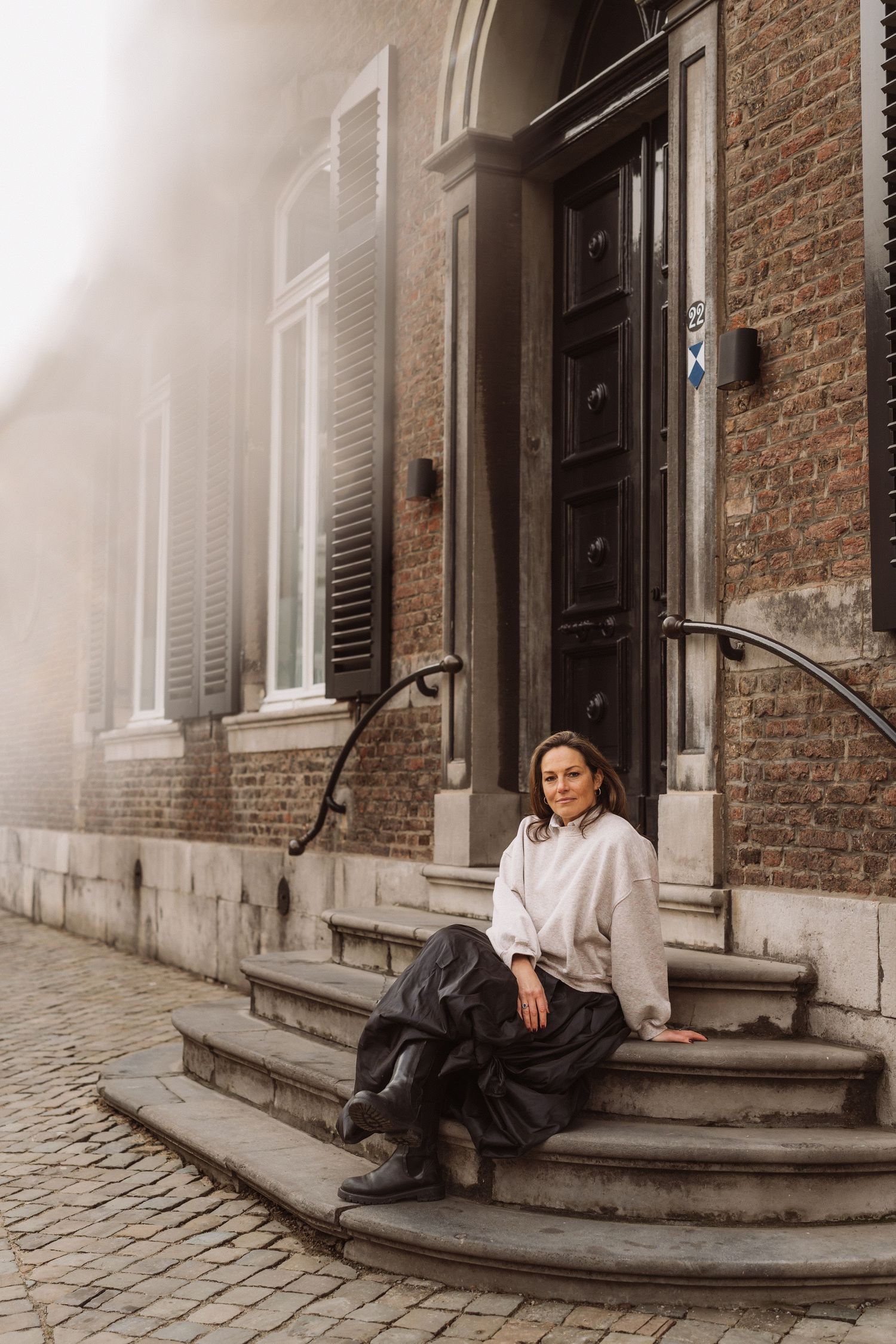 A person in casual attire sits on stone steps outside a historic brick building with white shuttered windows on a foggy day.