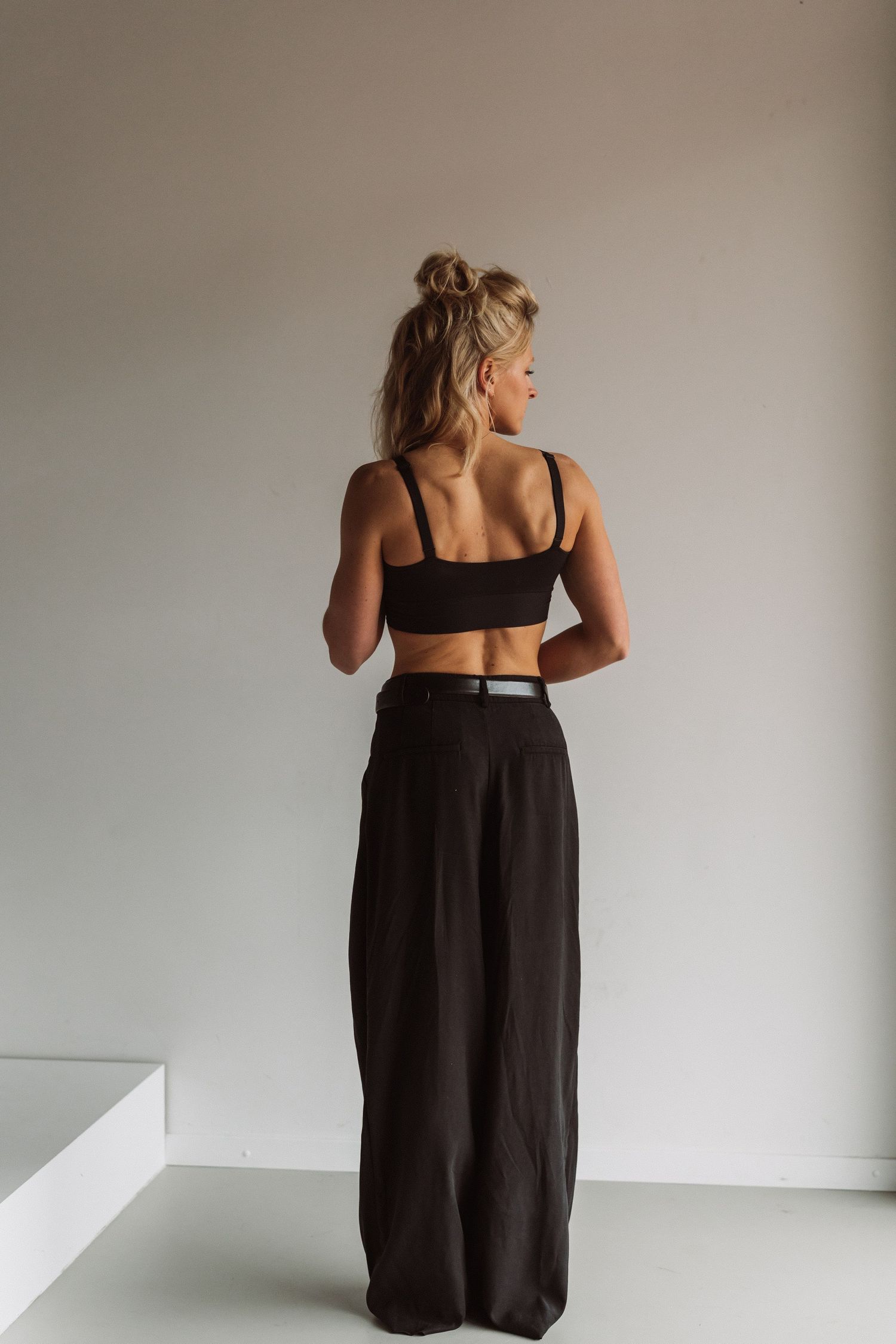 Woman in black crop top and wide-leg pants stands with back turned against white wall in minimalist setting.