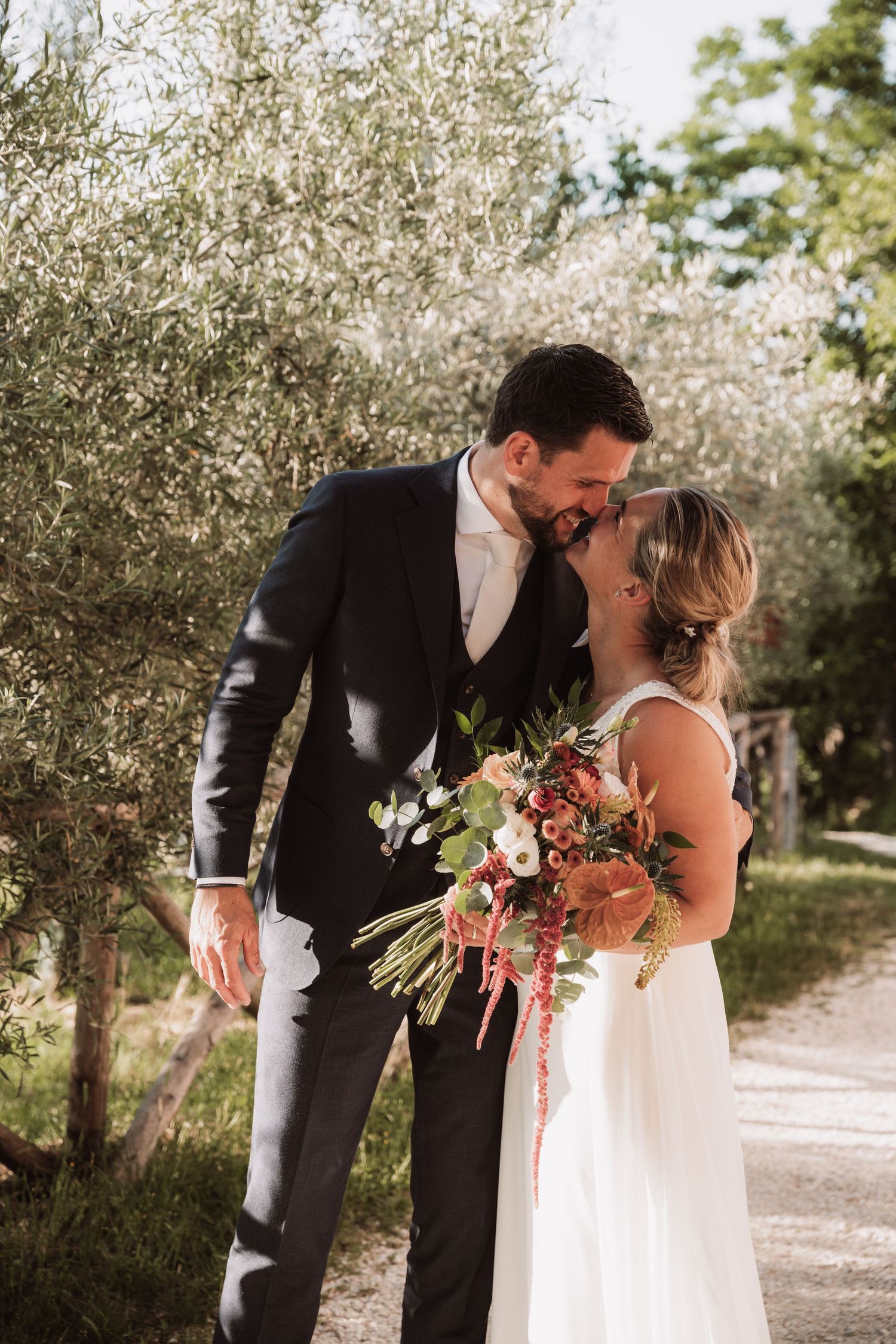 Romantic wedding couple sharing an intimate moment on a garden path surrounded by olive trees and holding a coral bouquet.