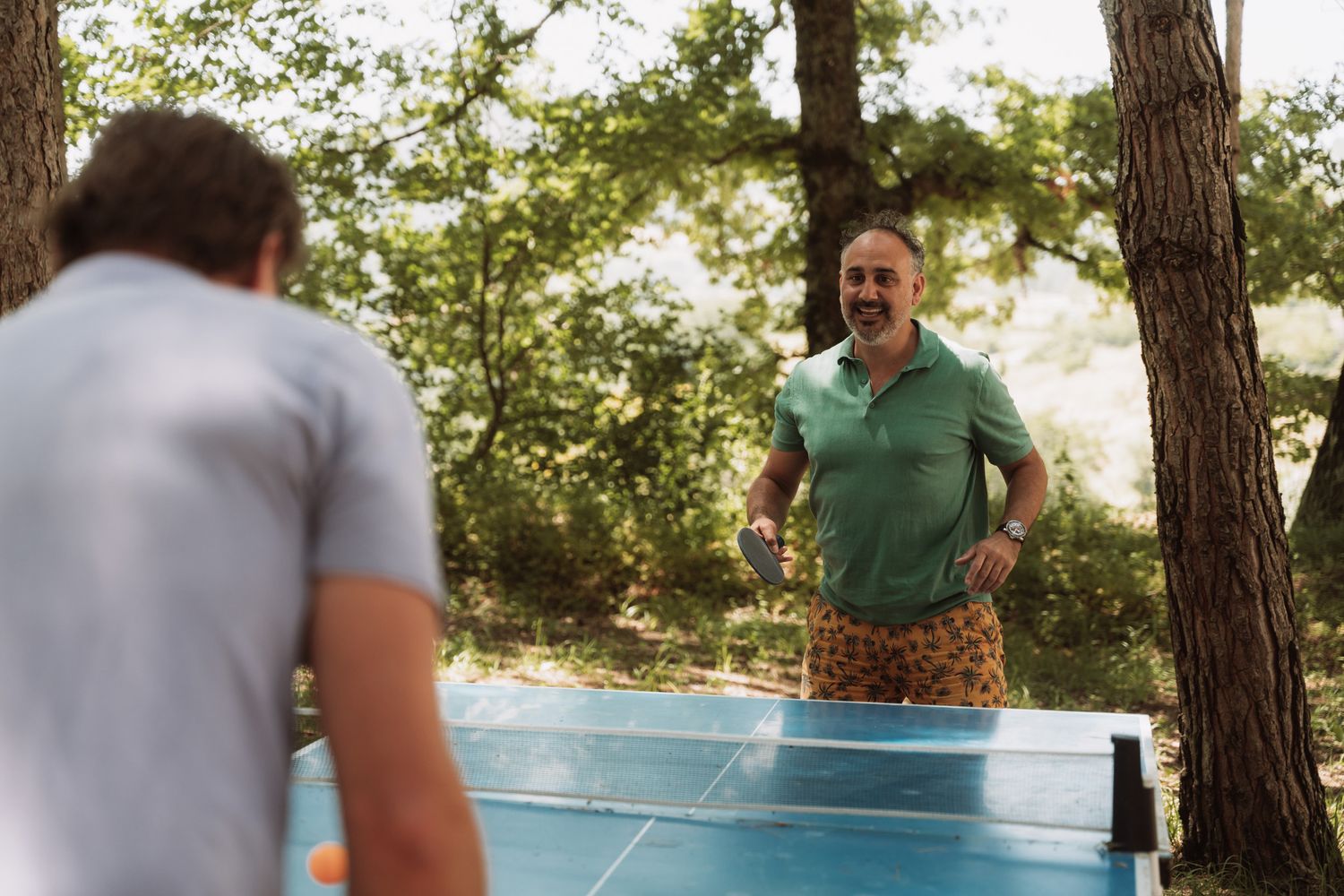 A person plays table tennis outdoors on a blue ping pong table surrounded by trees in a recreational setting.