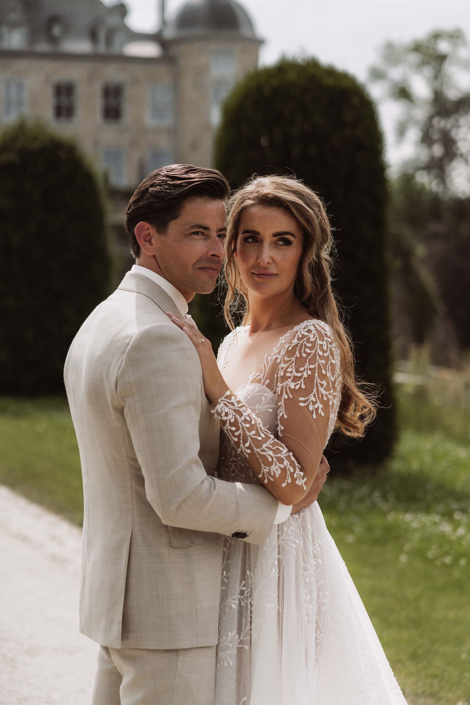 A romantic wedding couple poses outdoors in front of a chateau, with the bride in lace and groom in beige suit.