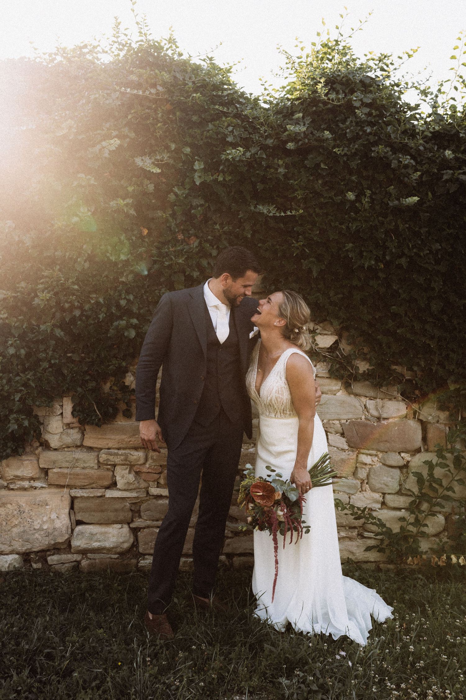 A romantic wedding couple embraces against a rustic stone wall at sunset in a garden setting.
