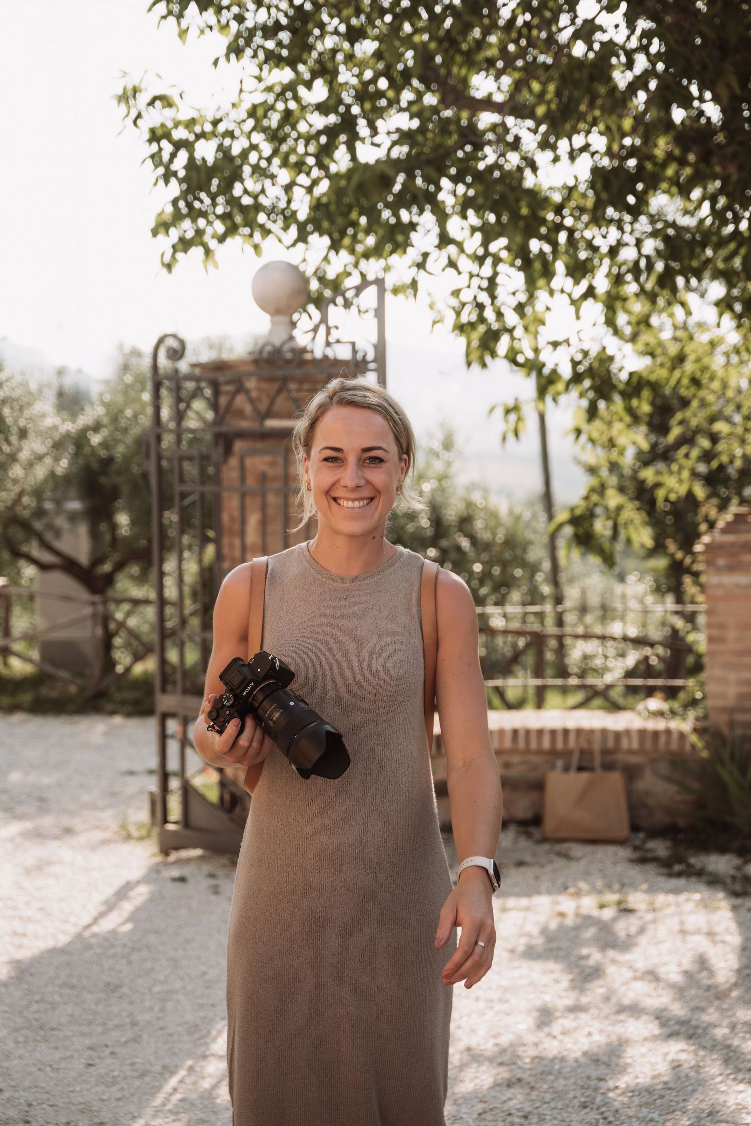 Person in a sleeveless beige dress holds a camera while standing outdoors on a sunny day among trees and brick walls.