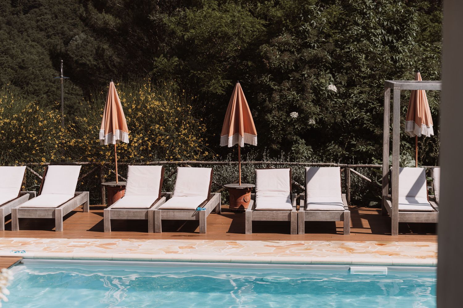Row of white lounge chairs with orange umbrellas arranged along the edge of a swimming pool on a wooden deck.
