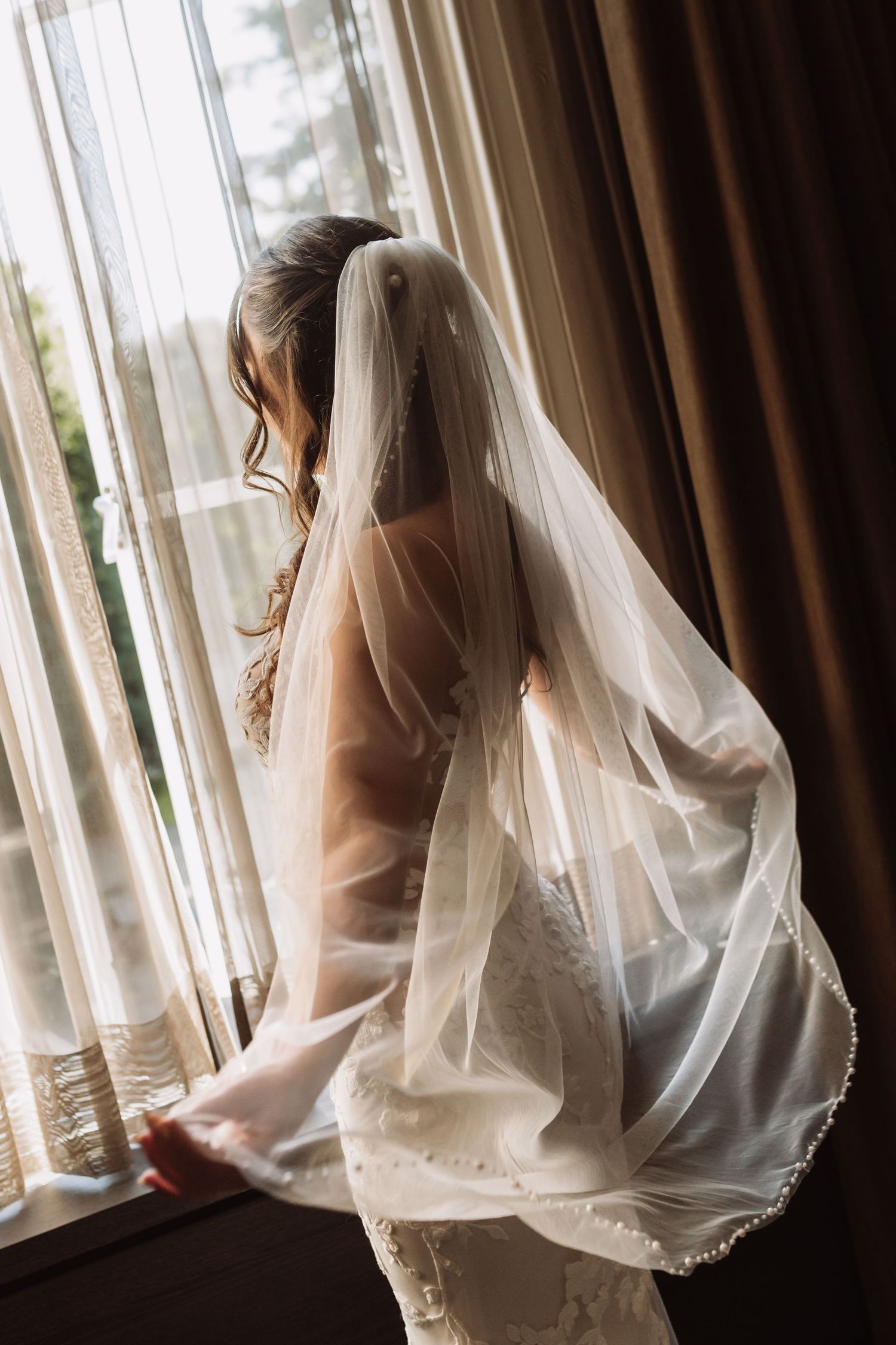 A bride in a flowing white veil gazes through sheer curtains in a series of artistic wedding day portraits.