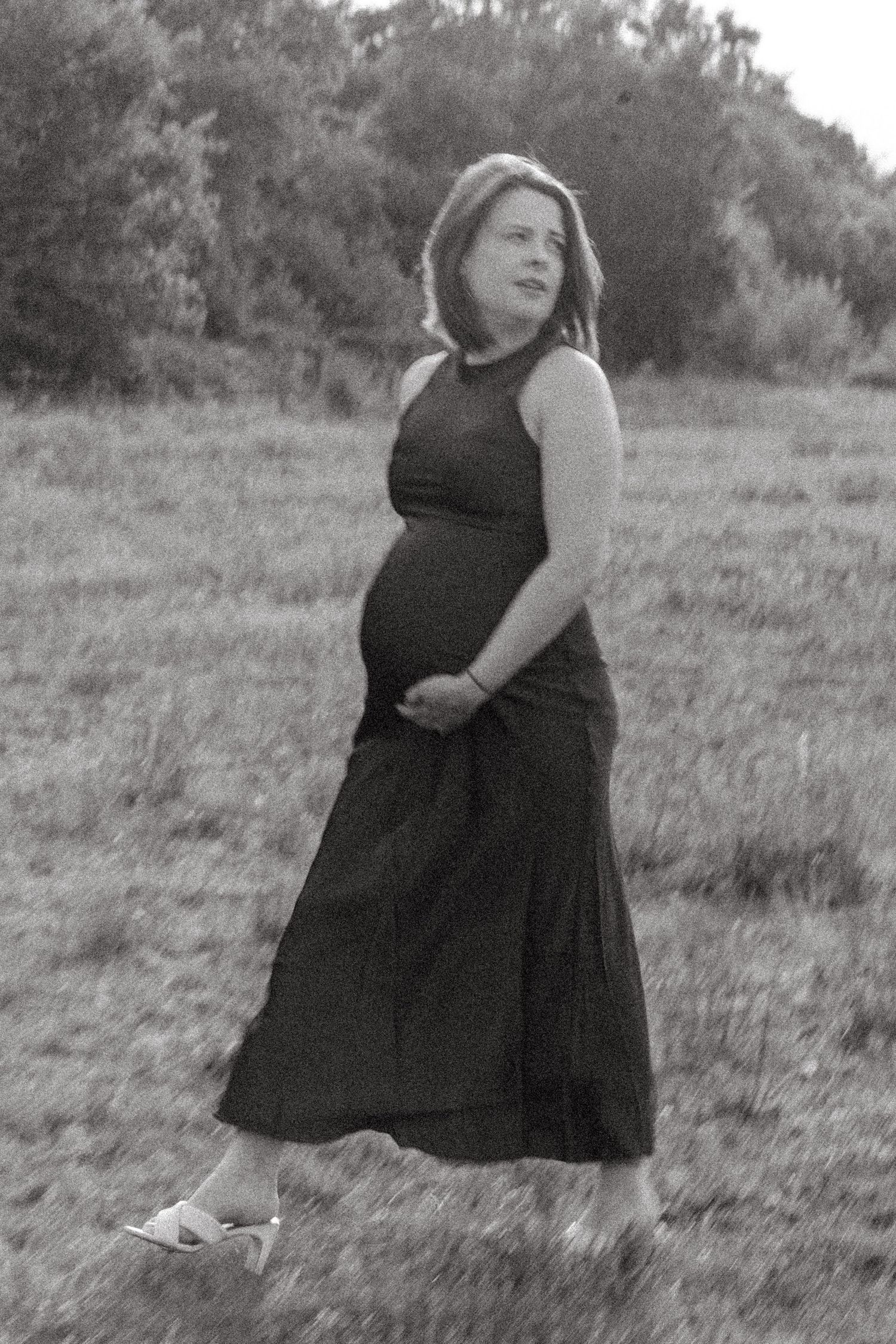 A figure in a black dress poses gracefully in a sunlit meadow with natural surroundings.