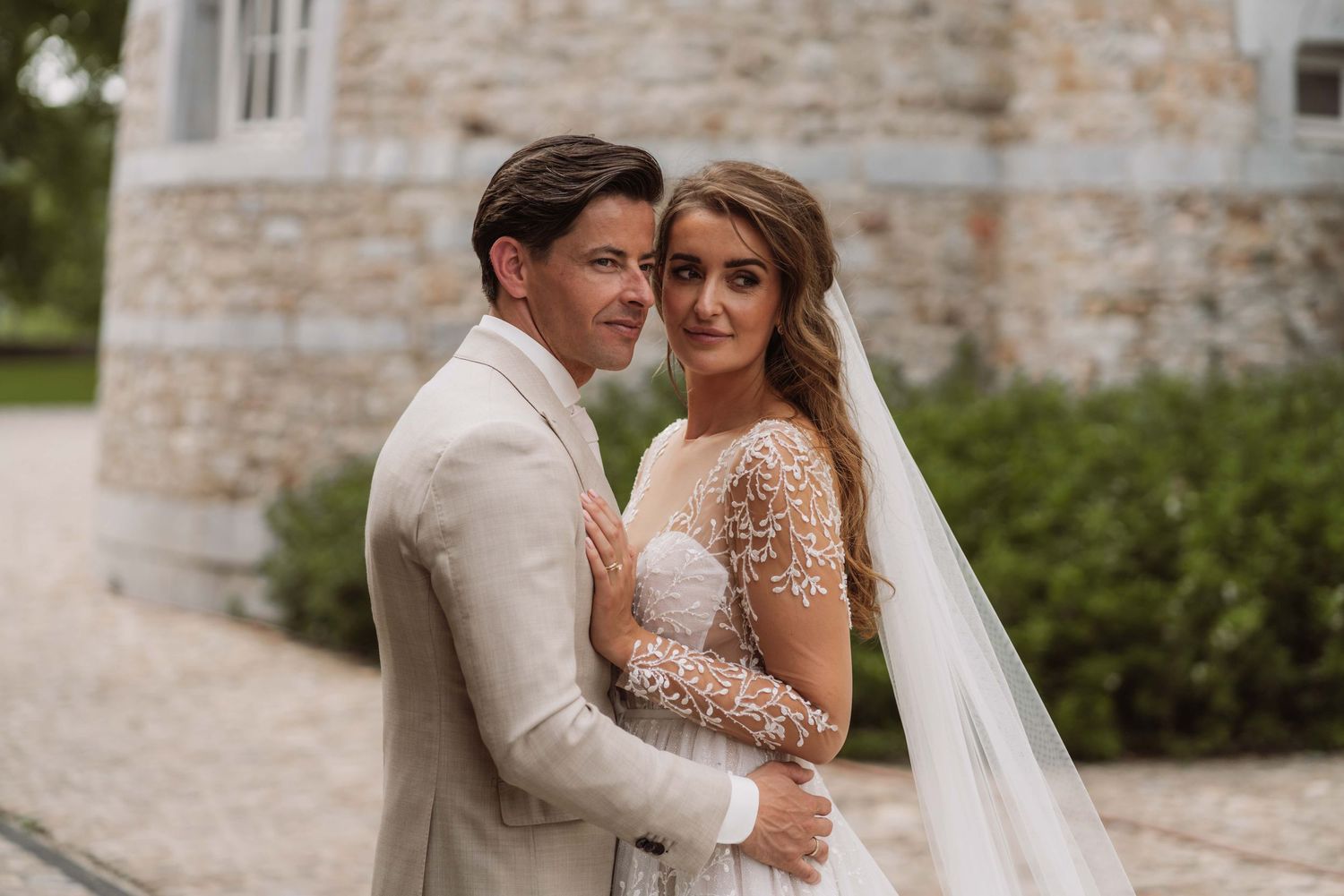A romantic moment between a couple in formal wedding attire against a stone wall backdrop.