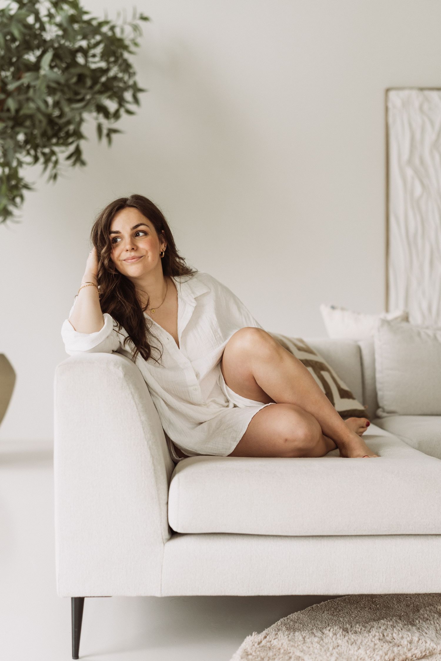 A figure in a white button-down shirt reclines on a light-colored sofa in a minimalist room with greenery.