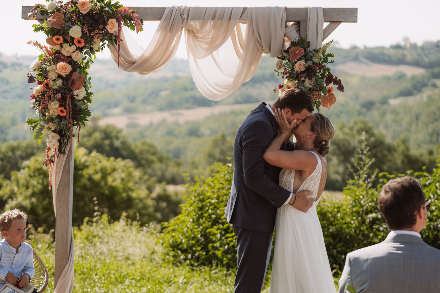 A romantic outdoor wedding ceremony overlooking rolling hills with floral arch decorations and flowing blush drapery.