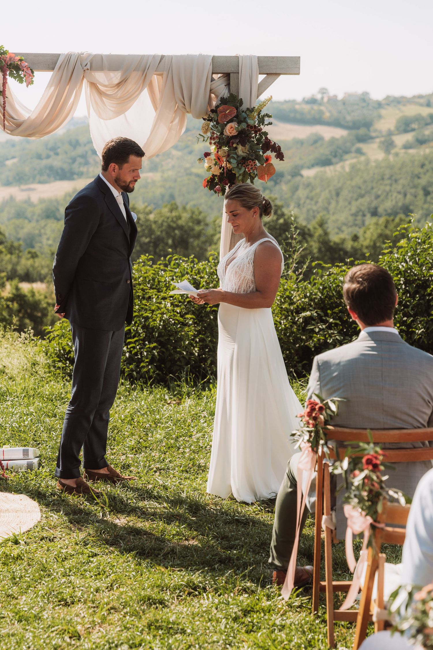 An intimate outdoor wedding ceremony takes place on a hilltop with draped fabric and floral arrangements against a scenic view.