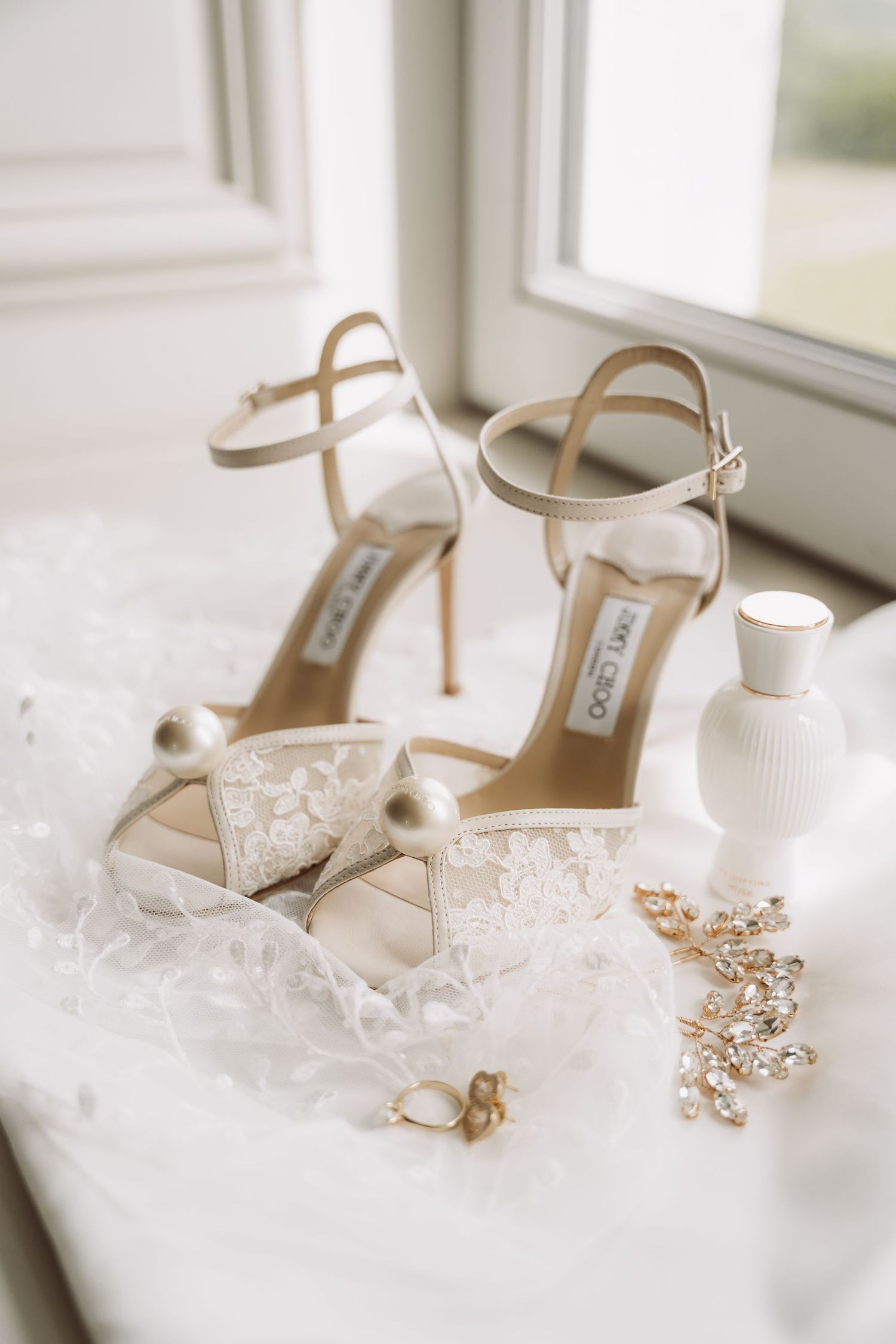 Elegant ivory wedding shoes with crystal embellishments displayed on white fabric near a perfume bottle and jewelry.
