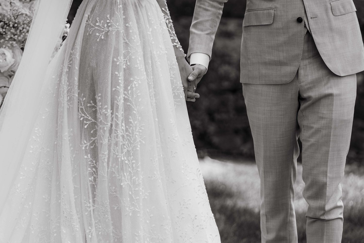 Black and white close-up of couple holding hands at wedding, showing detailed lace dress and gray suit.