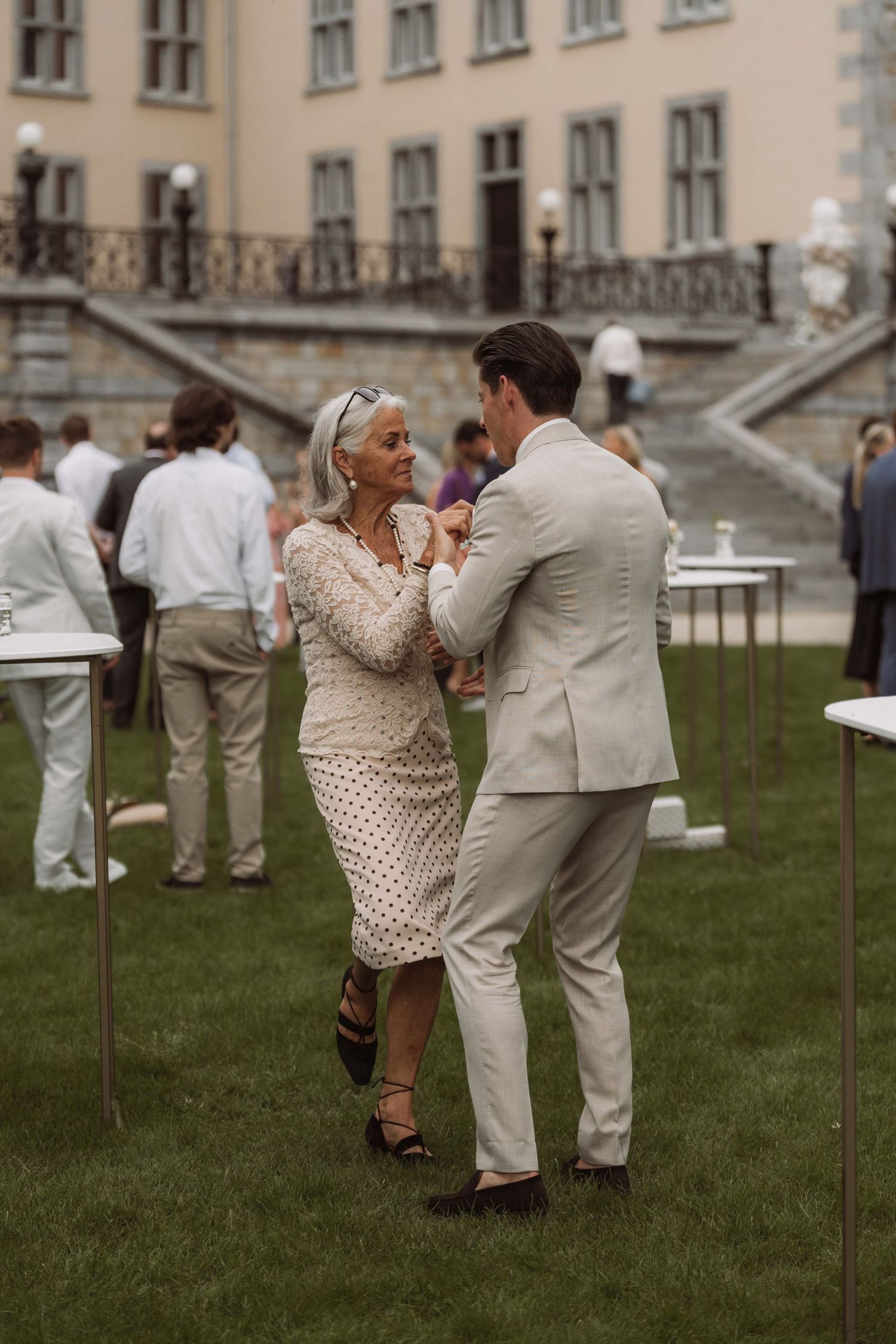 Couples in white and beige attire dance together on a grassy lawn outside an elegant building.