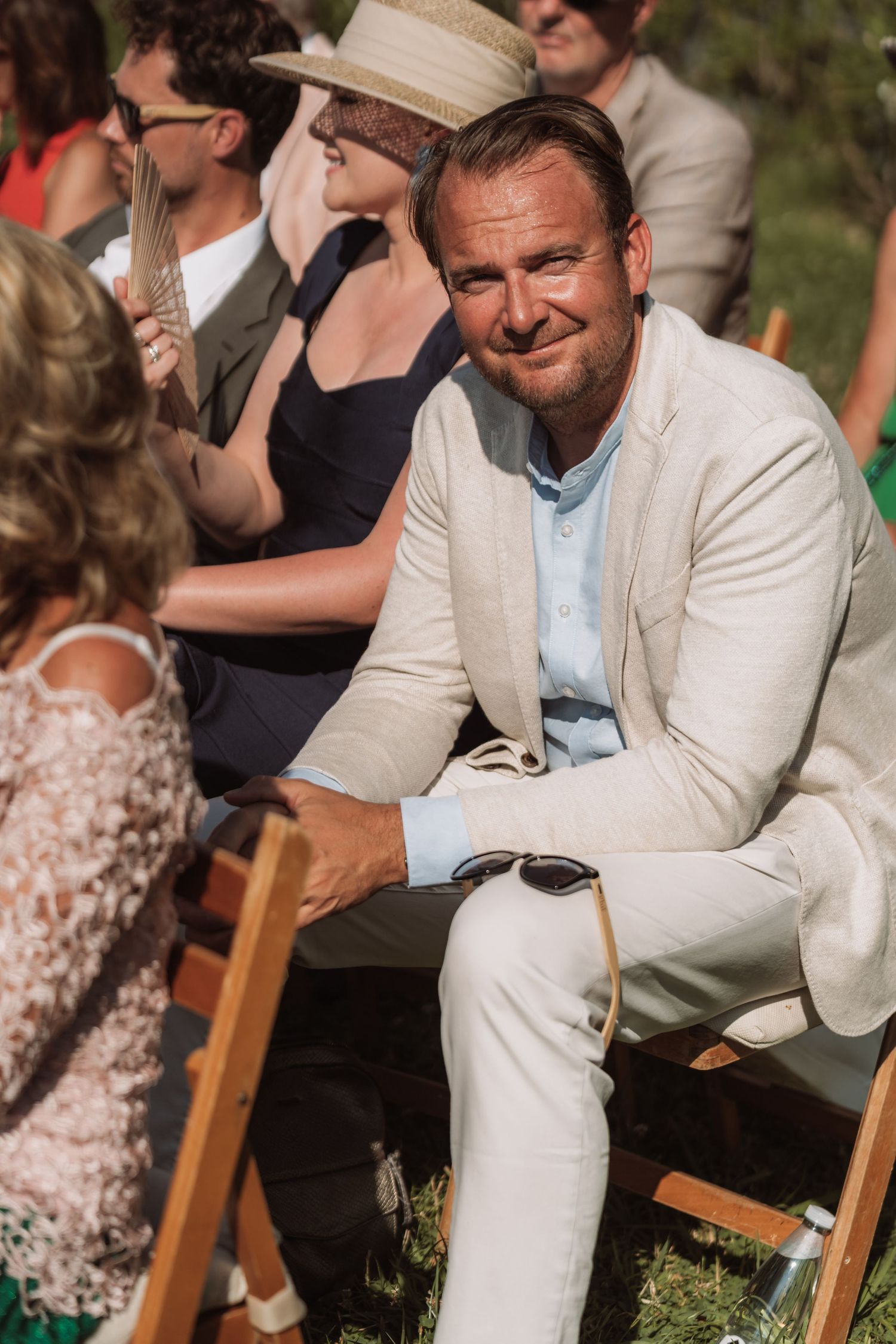 A person in a light colored suit jacket sits casually on a wooden chair outdoors during what appears to be an event.