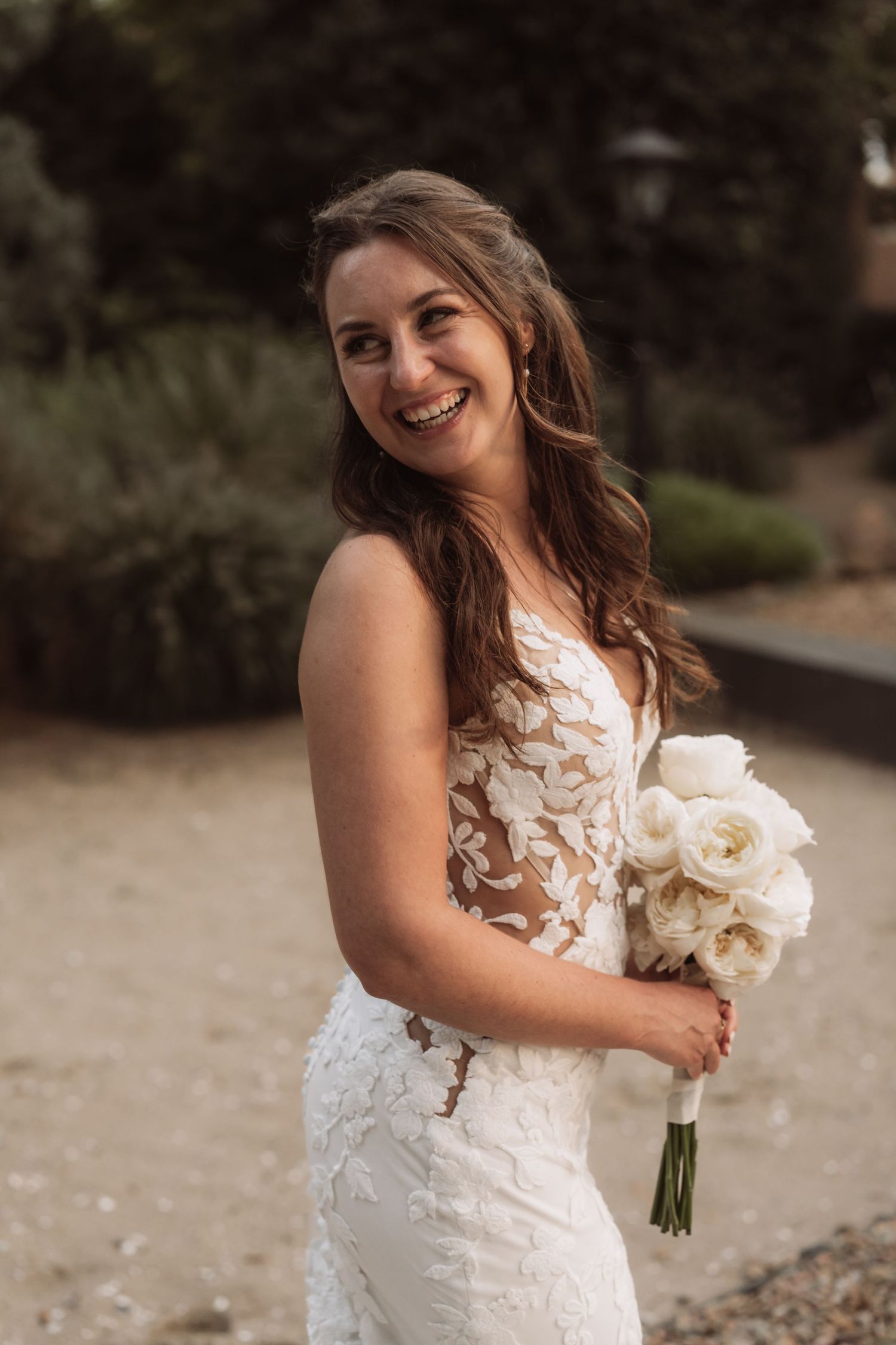 A bride in a white lace wedding dress holds a bouquet of cream-colored flowers while posing outdoors.