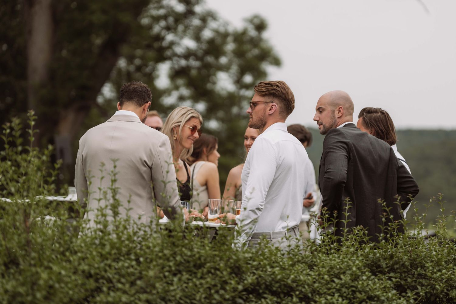 Wedding guests mingle and socialize outdoors during a garden reception with lush greenery in the background.