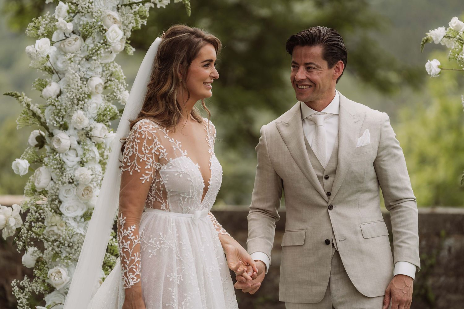 A couple in wedding attire stands together under white floral arches in a lush garden setting.