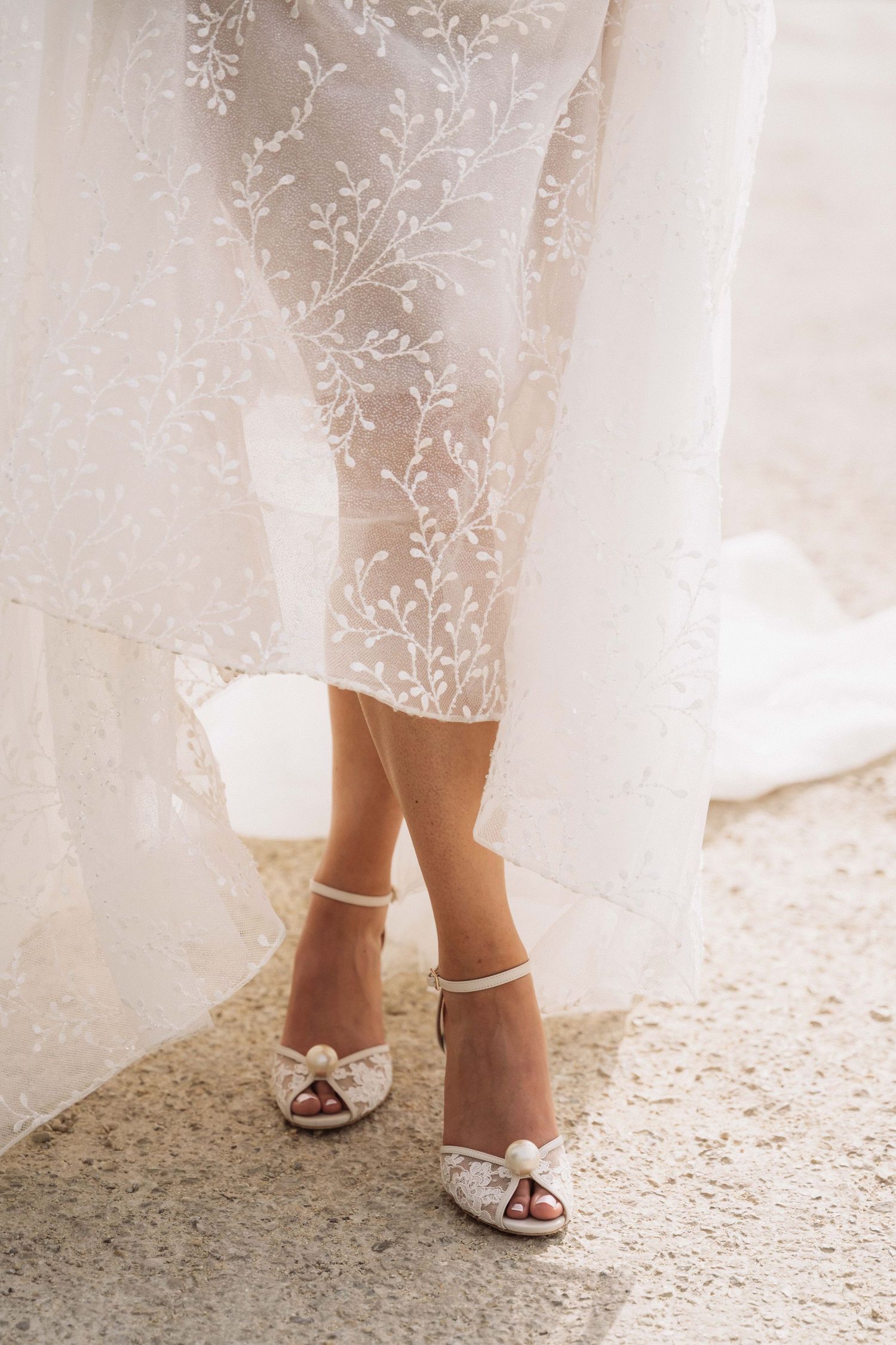 A bride's feet in elegant white peep-toe wedding shoes peek out from beneath a delicate lace wedding gown.