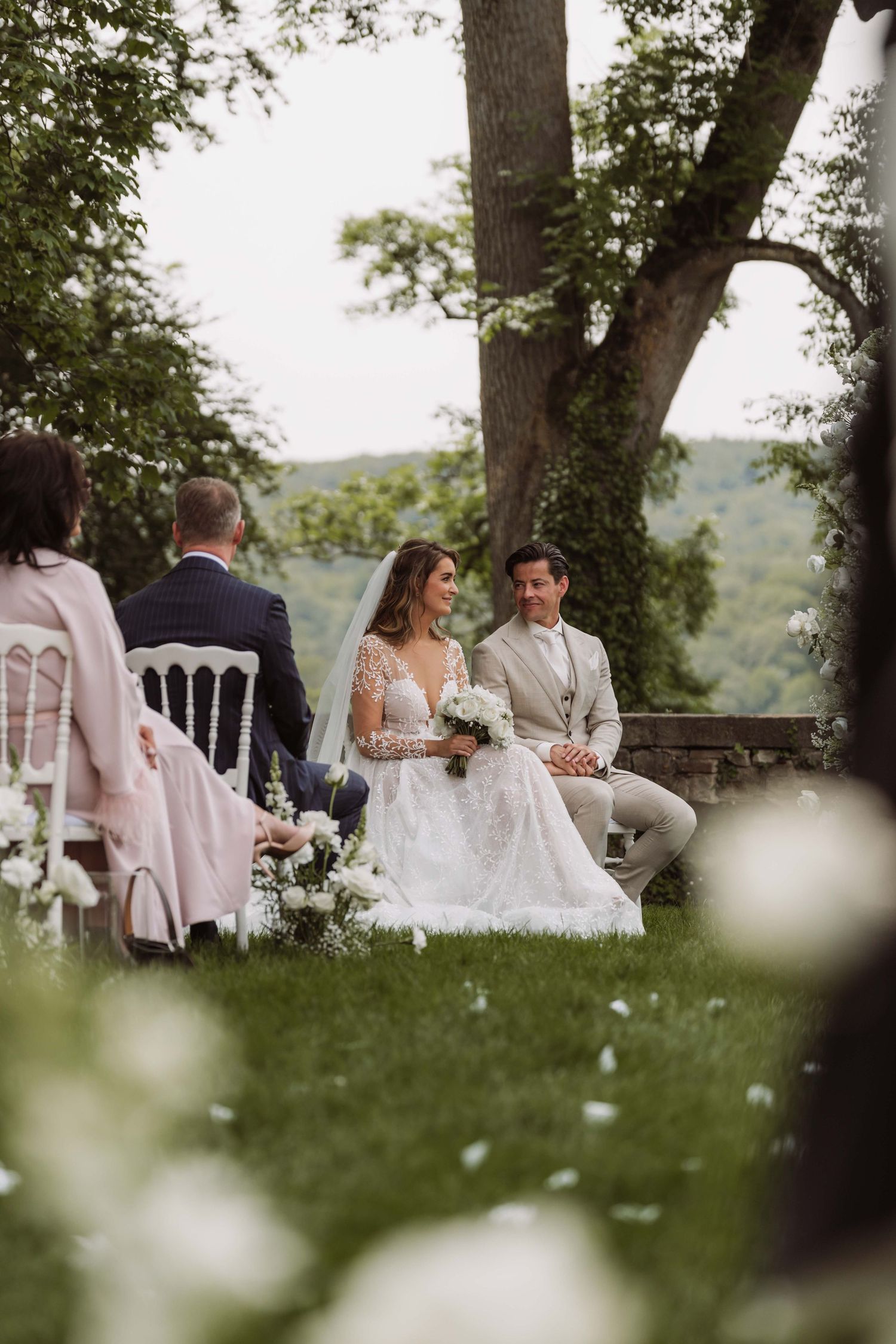 Wedding guests relax on the grass during an outdoor summer reception beneath towering trees.