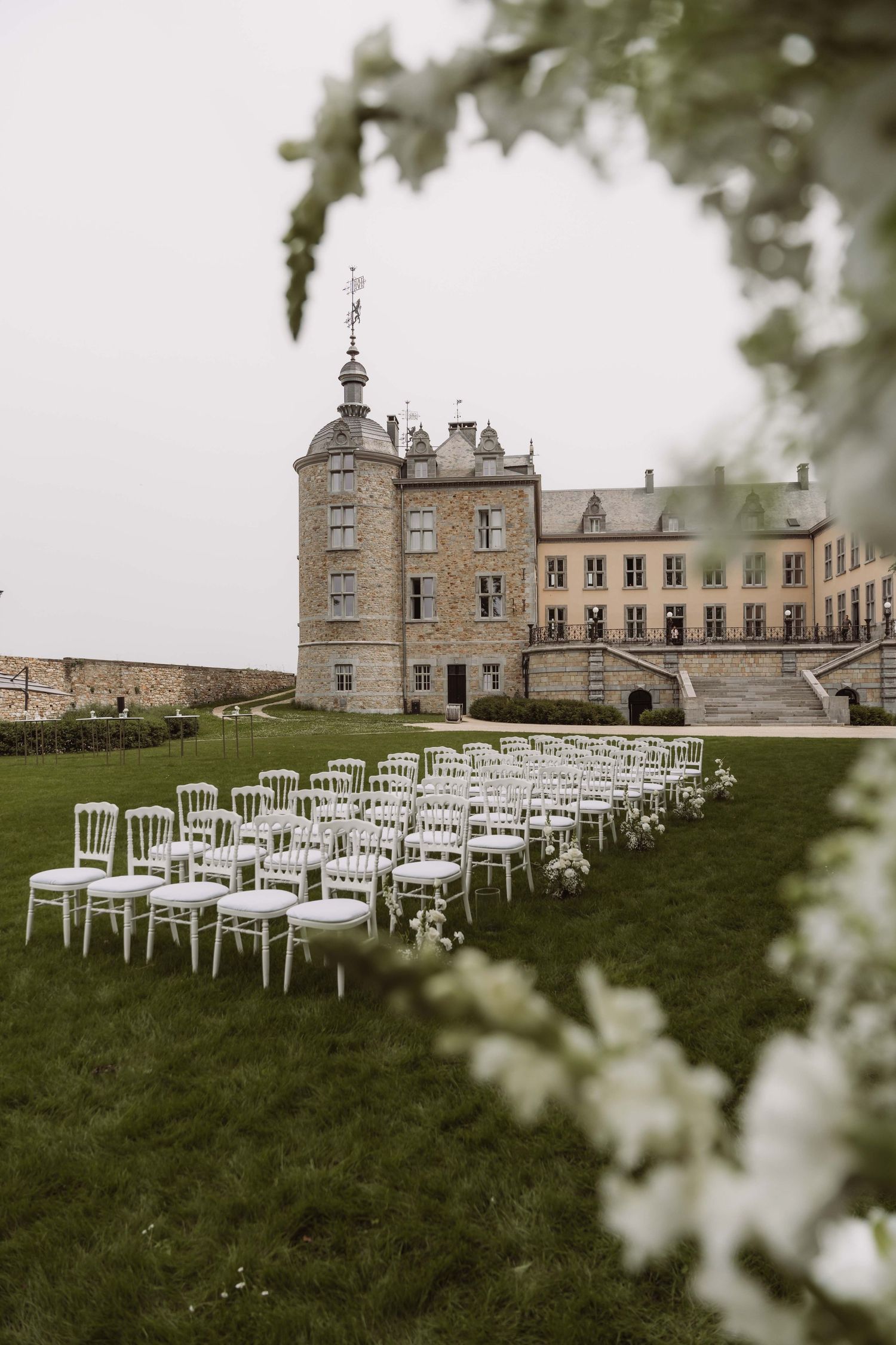 White chairs arranged for an outdoor wedding ceremony in front of a historic stone castle with blooming flowers in foreground.
