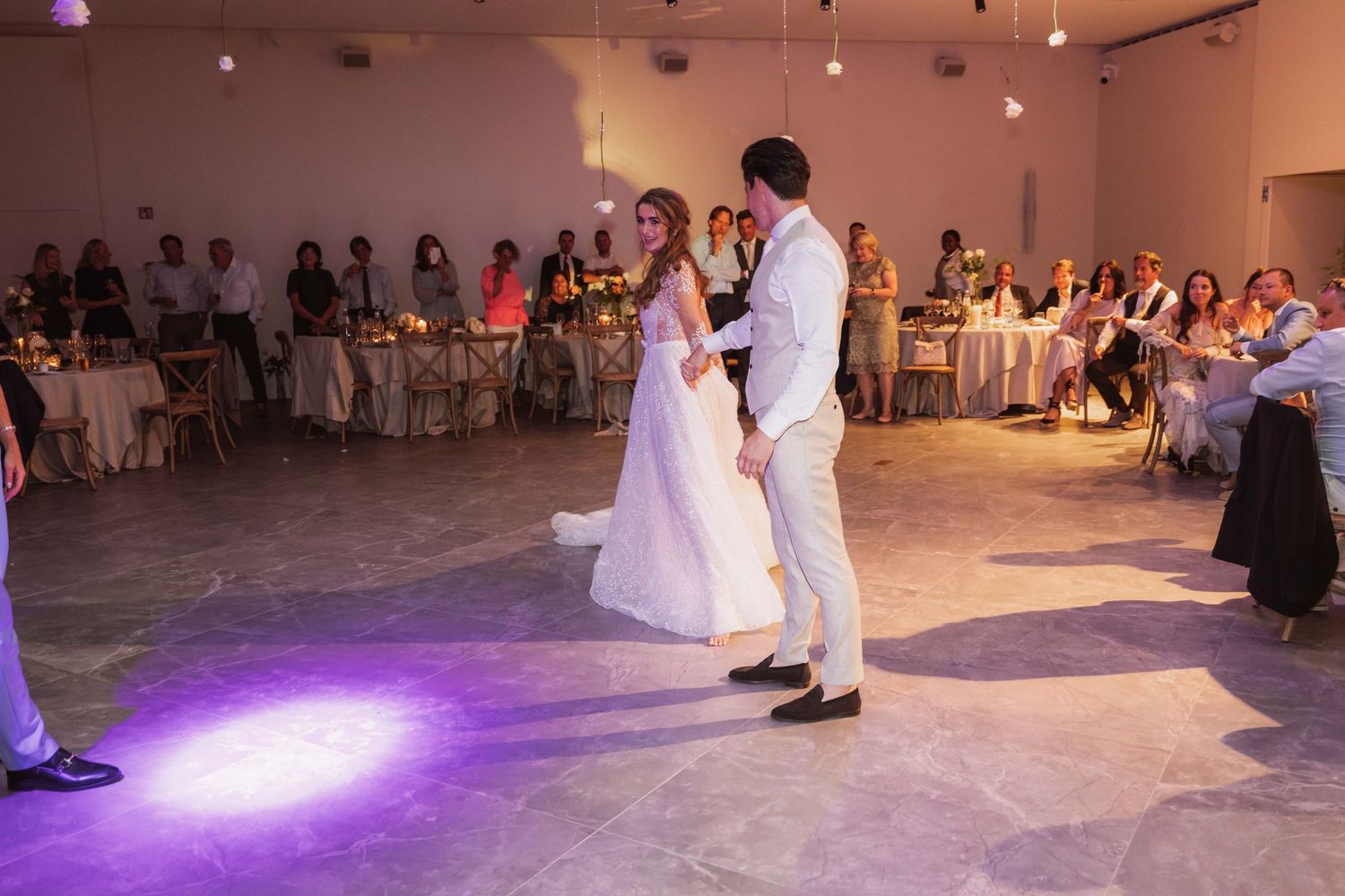 Wedding reception guests dance and celebrate on the dance floor under purple mood lighting at an indoor venue.