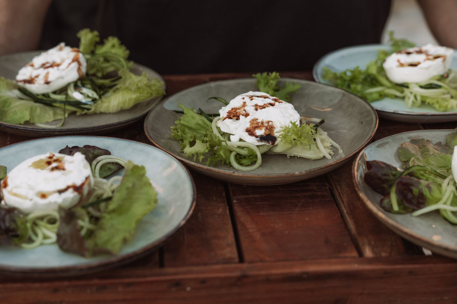 Fresh salad plates with poached eggs, leafy greens and garnishes arranged in a row on a wooden surface.