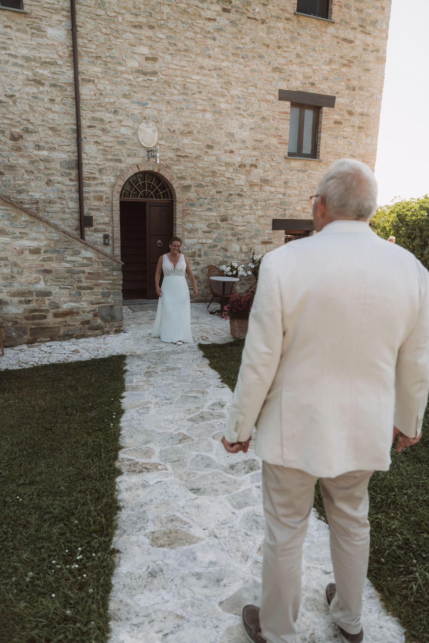 Two people in cream-colored formal attire walk along a stone path beside a grassy lawn and stone building wall.