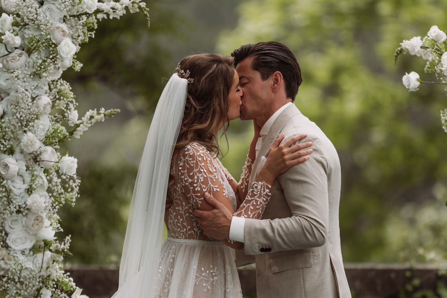 A romantic wedding couple shares multiple tender kisses under a floral arch decorated with white blossoms.
