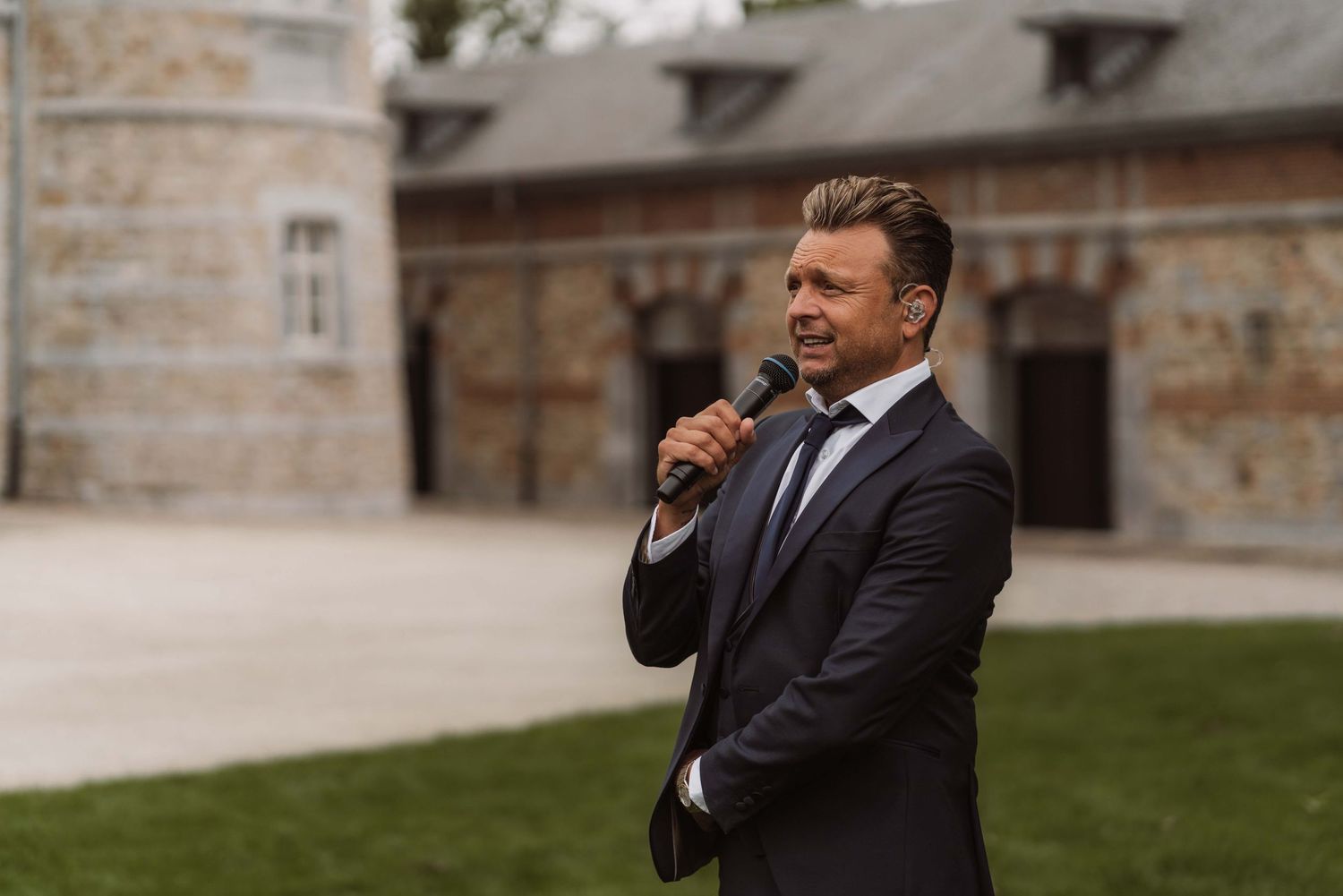 A person in a dark suit adjusts their tie while standing outside a stone building on a cloudy day.