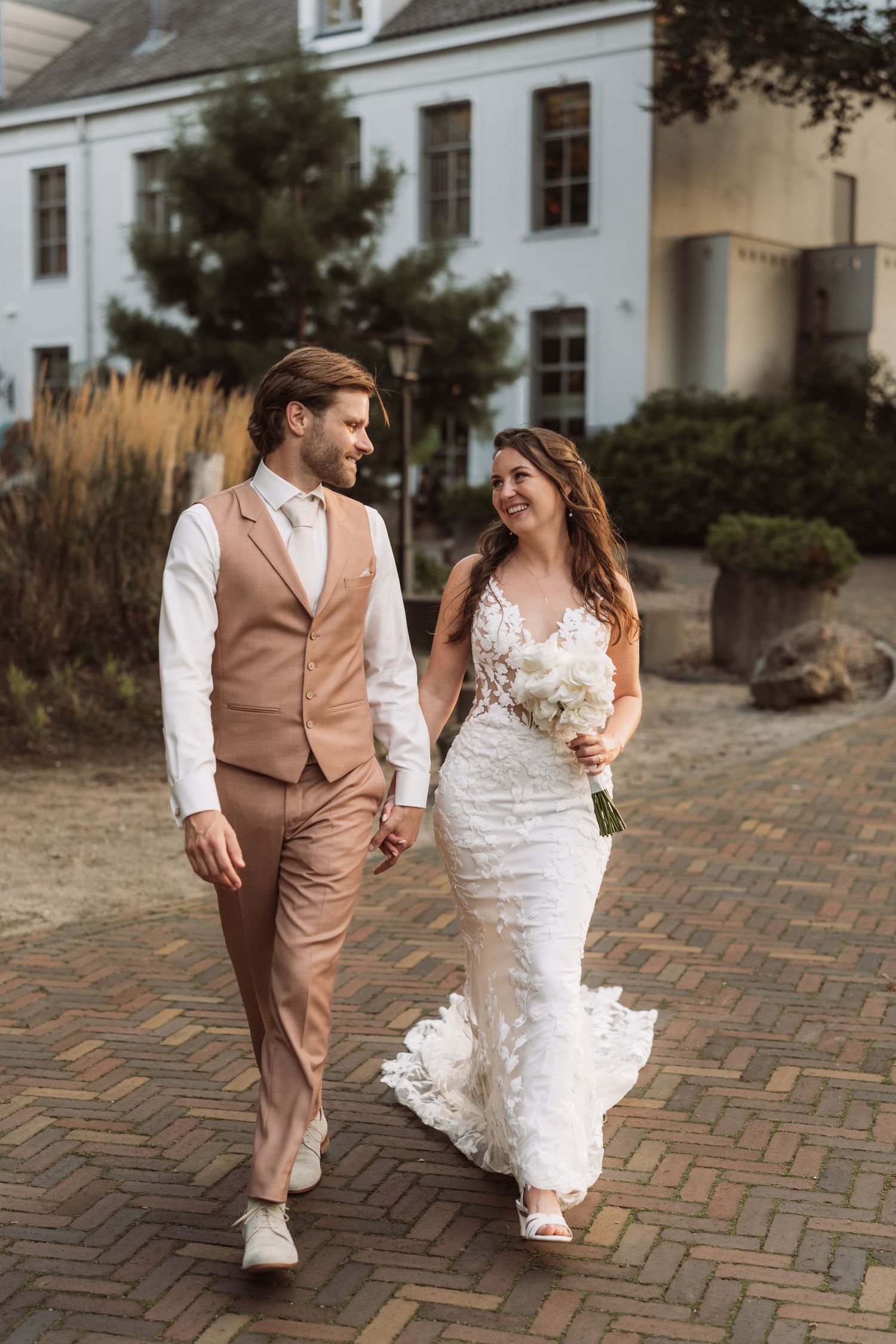 A couple in wedding attire walks together on a brick courtyard pathway at dusk.