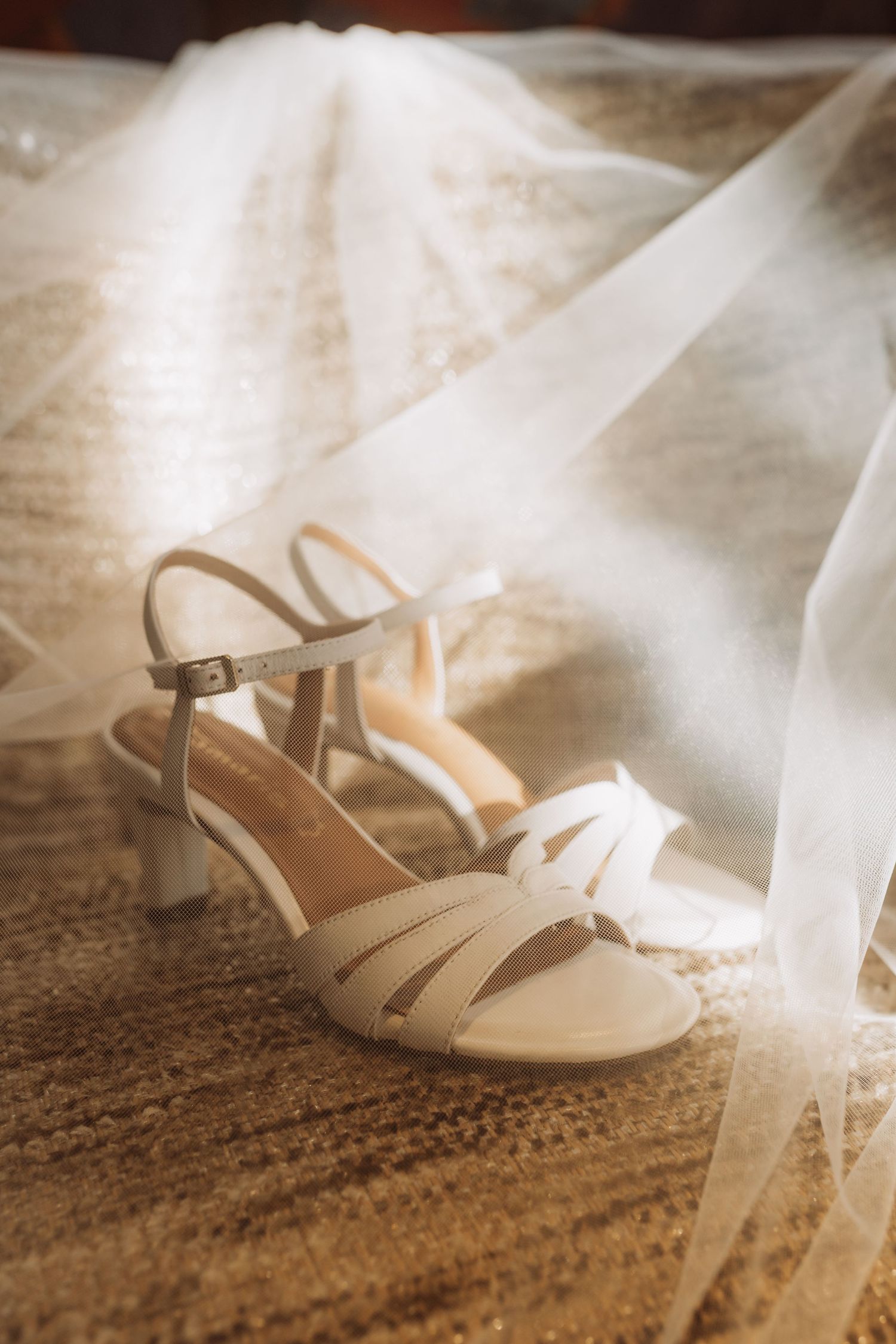 White wedding shoes with delicate straps rest beside flowing tulle fabric on a textured brown carpet in soft lighting.
