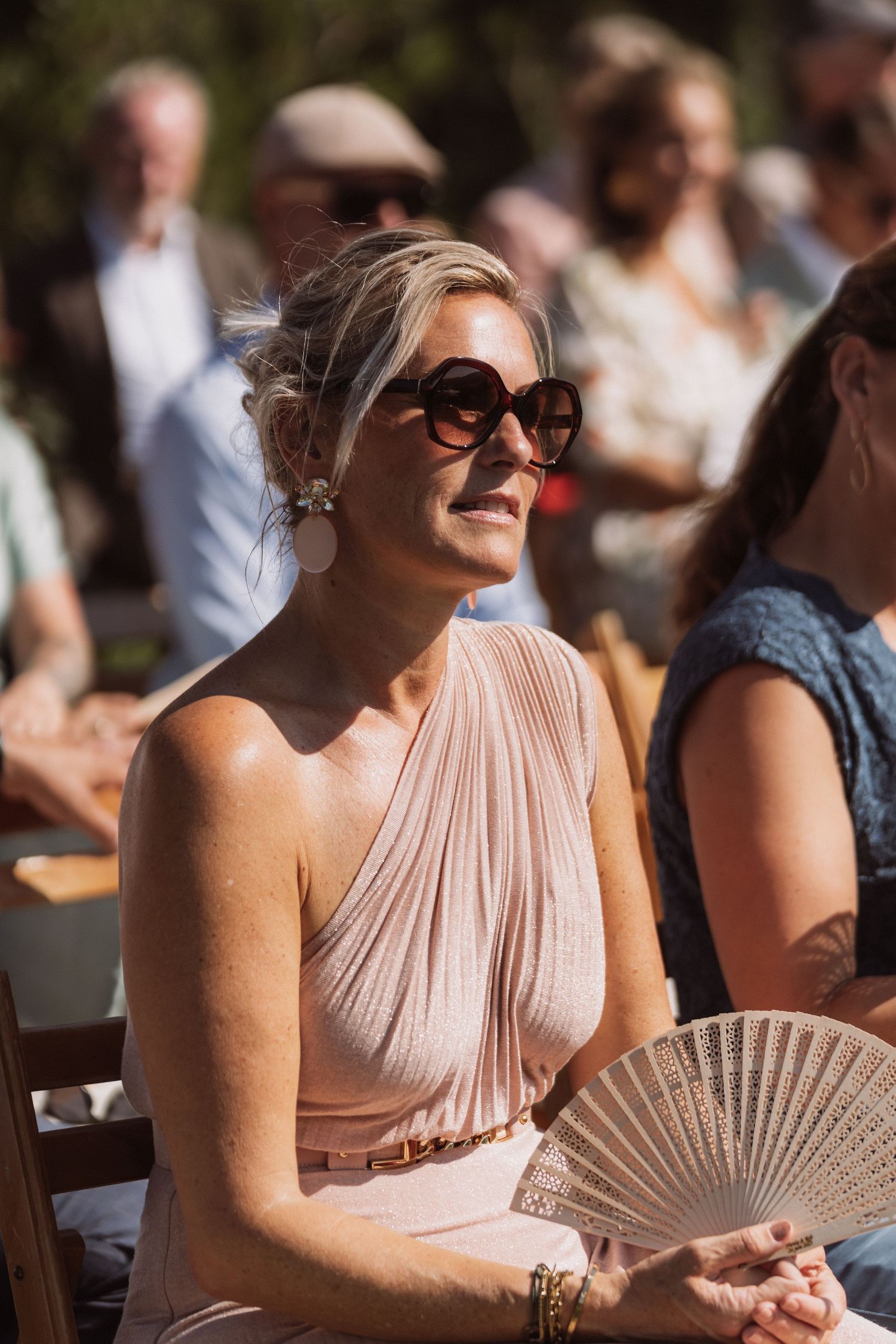 A wedding guest in a blush pink pleated dress holds a decorative fan while sitting outdoors at a summer ceremony.