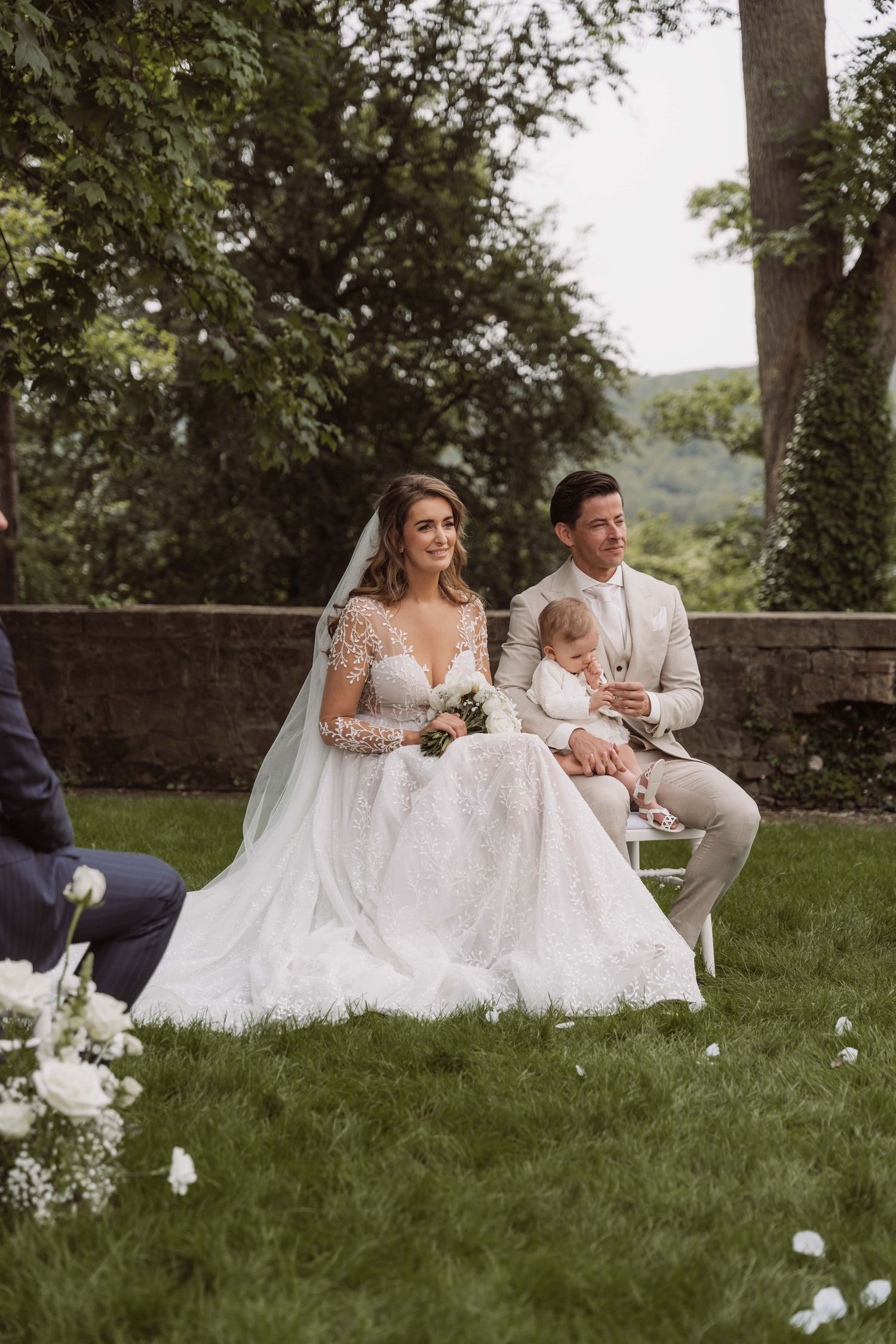 A couple and baby sit on chairs during an outdoor wedding ceremony with white floral decorations on a grassy lawn.