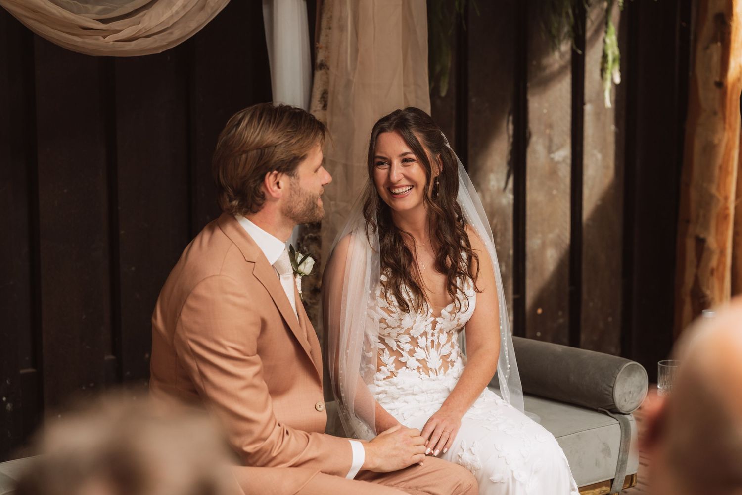 A couple shares a joyful moment together at their rustic wedding ceremony, with the bride in white lace and groom in tan suit.