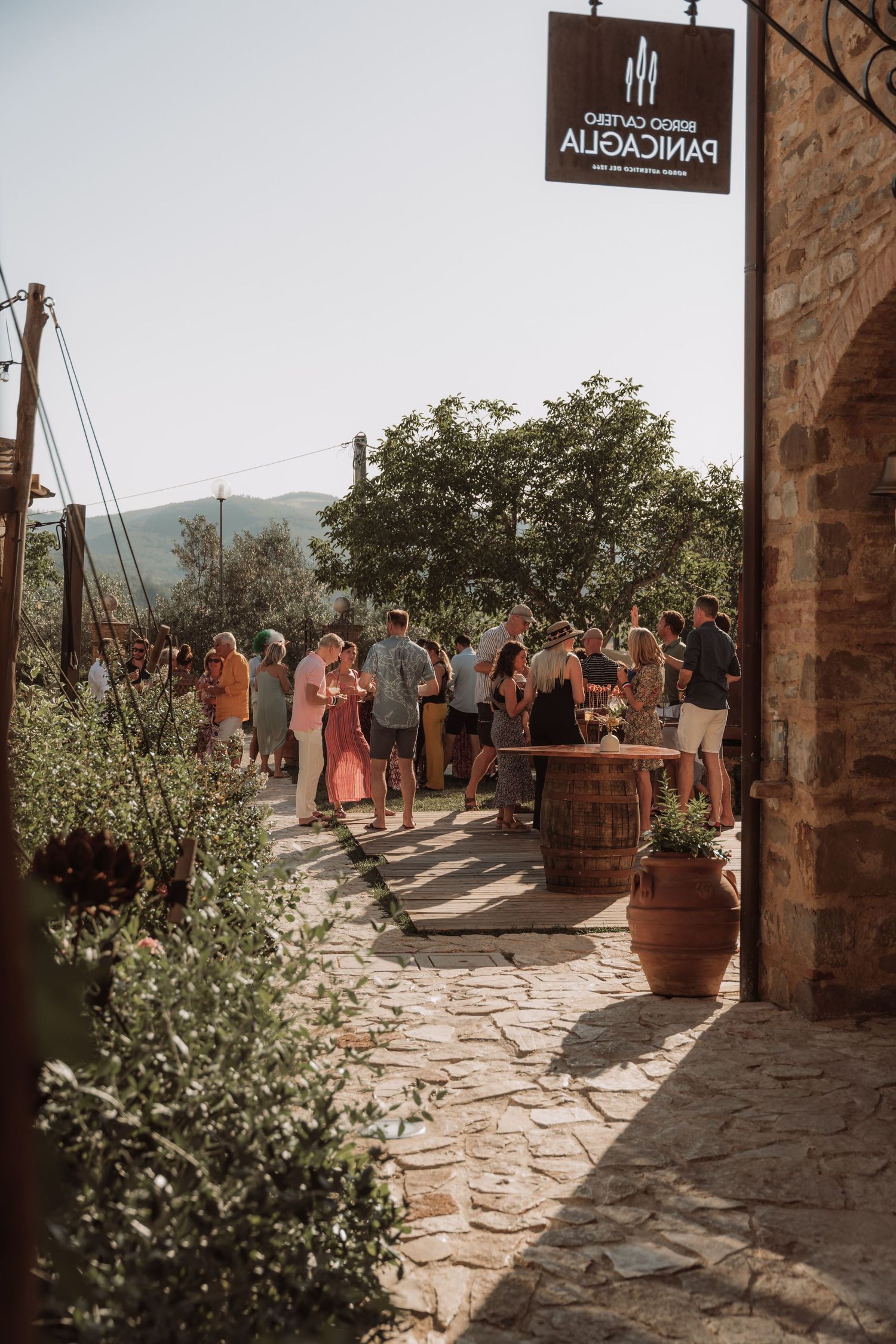 Guests gather on a rustic stone terrace with potted plants at an outdoor wedding venue in warm sunlight.
