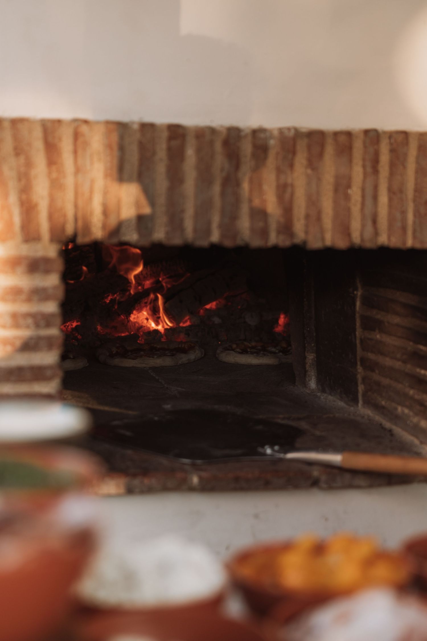 Glowing embers and flames burn inside a rustic brick pizza oven with blurred dishes in the foreground.