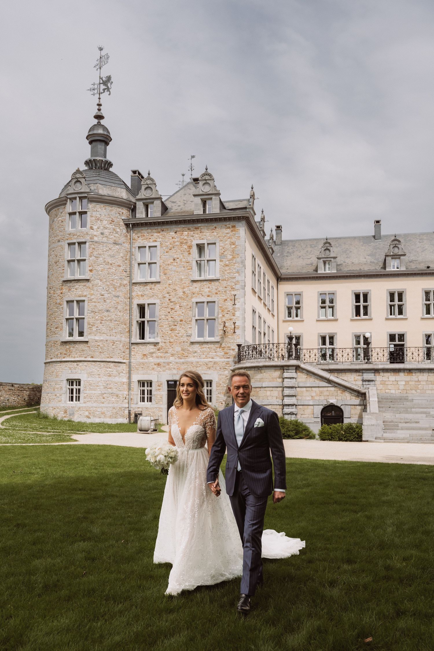 Newlyweds walking together on the lawn of a grand stone chateau on their wedding day.