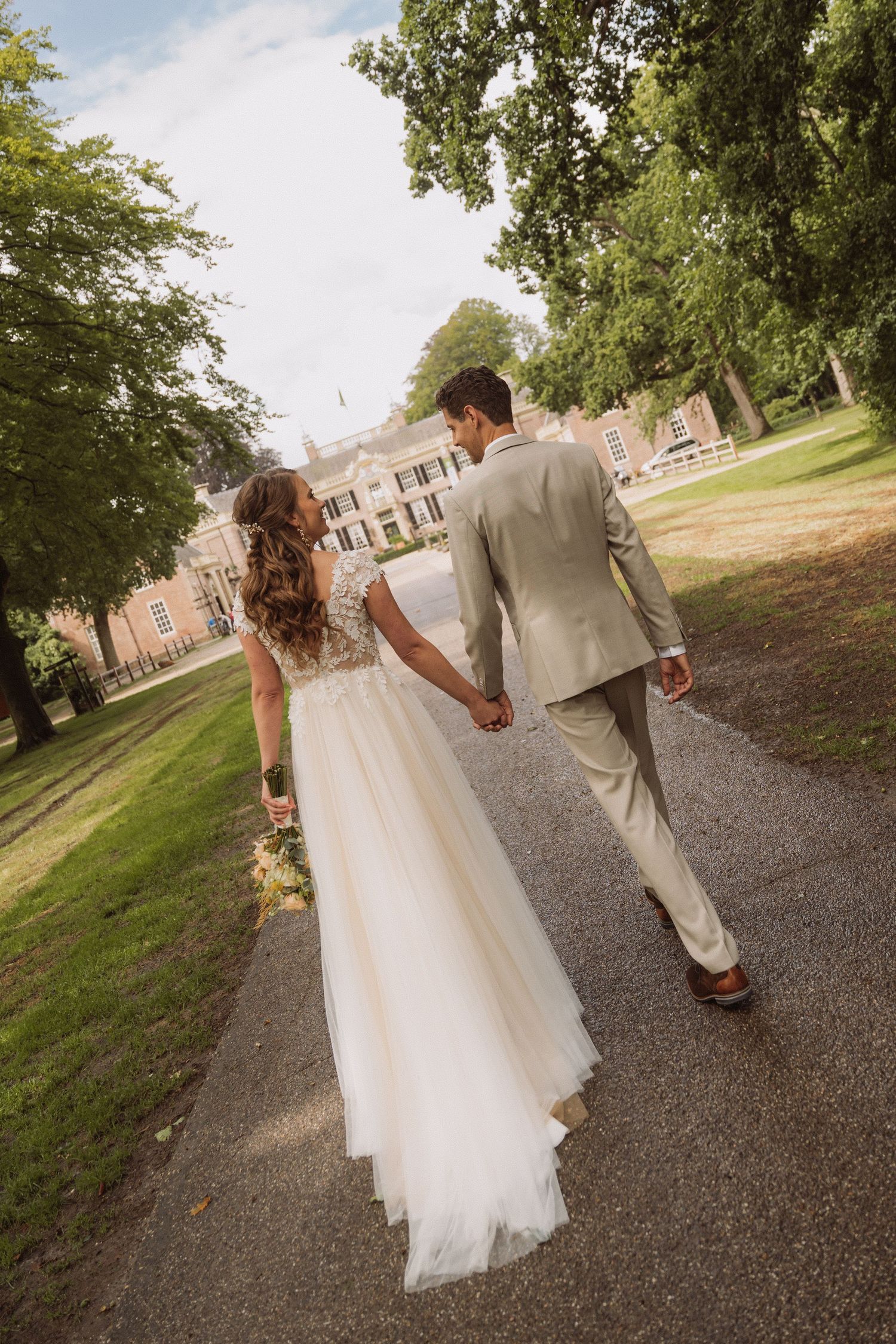 Wedding couple walking down a tree-lined path in formal attire.