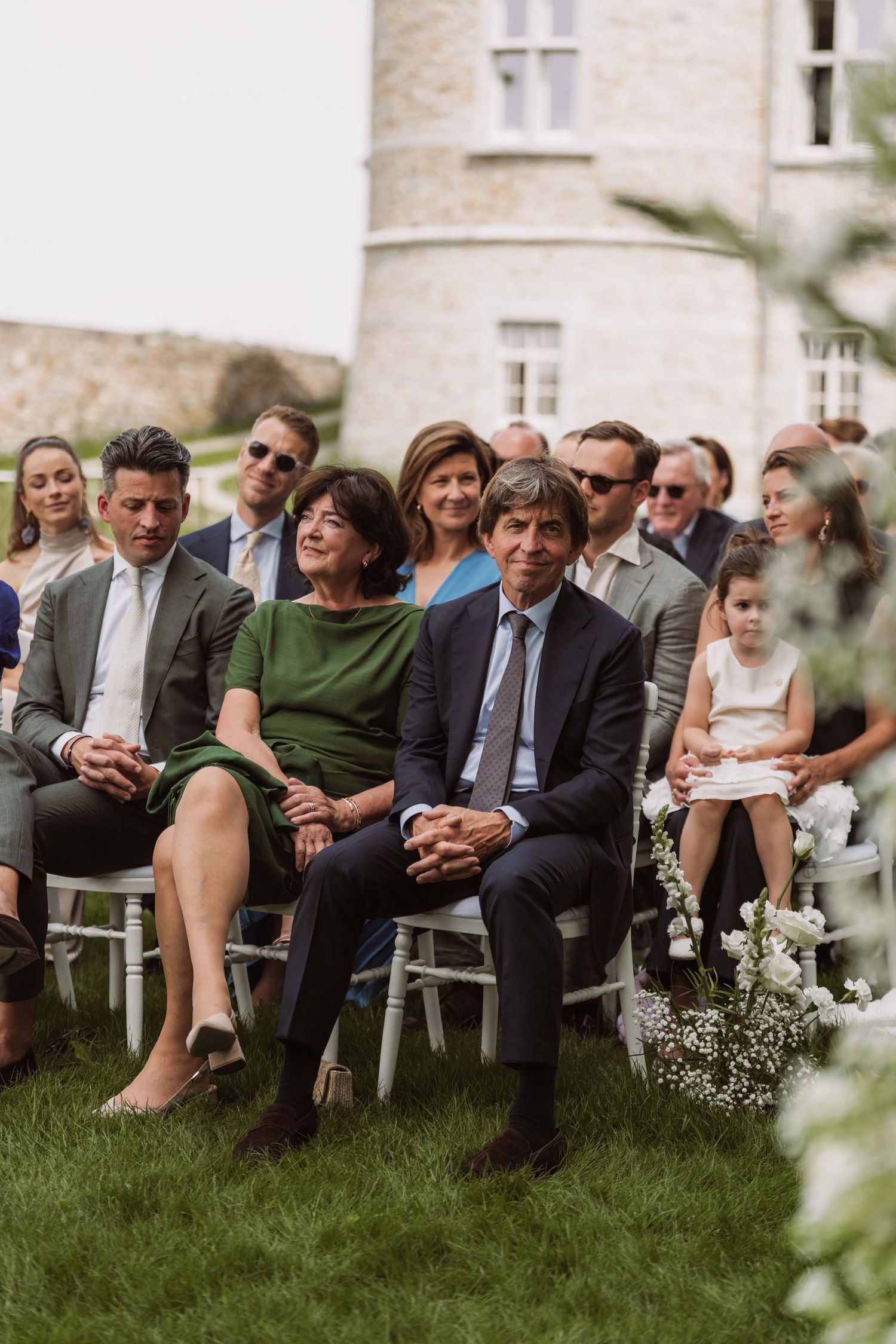 Wedding guests seated on white chairs on a lawn outside a stone building during an outdoor ceremony.