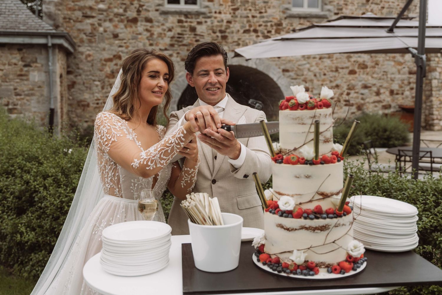 A couple in wedding attire laughs while cutting their white wedding cake decorated with fresh strawberries.