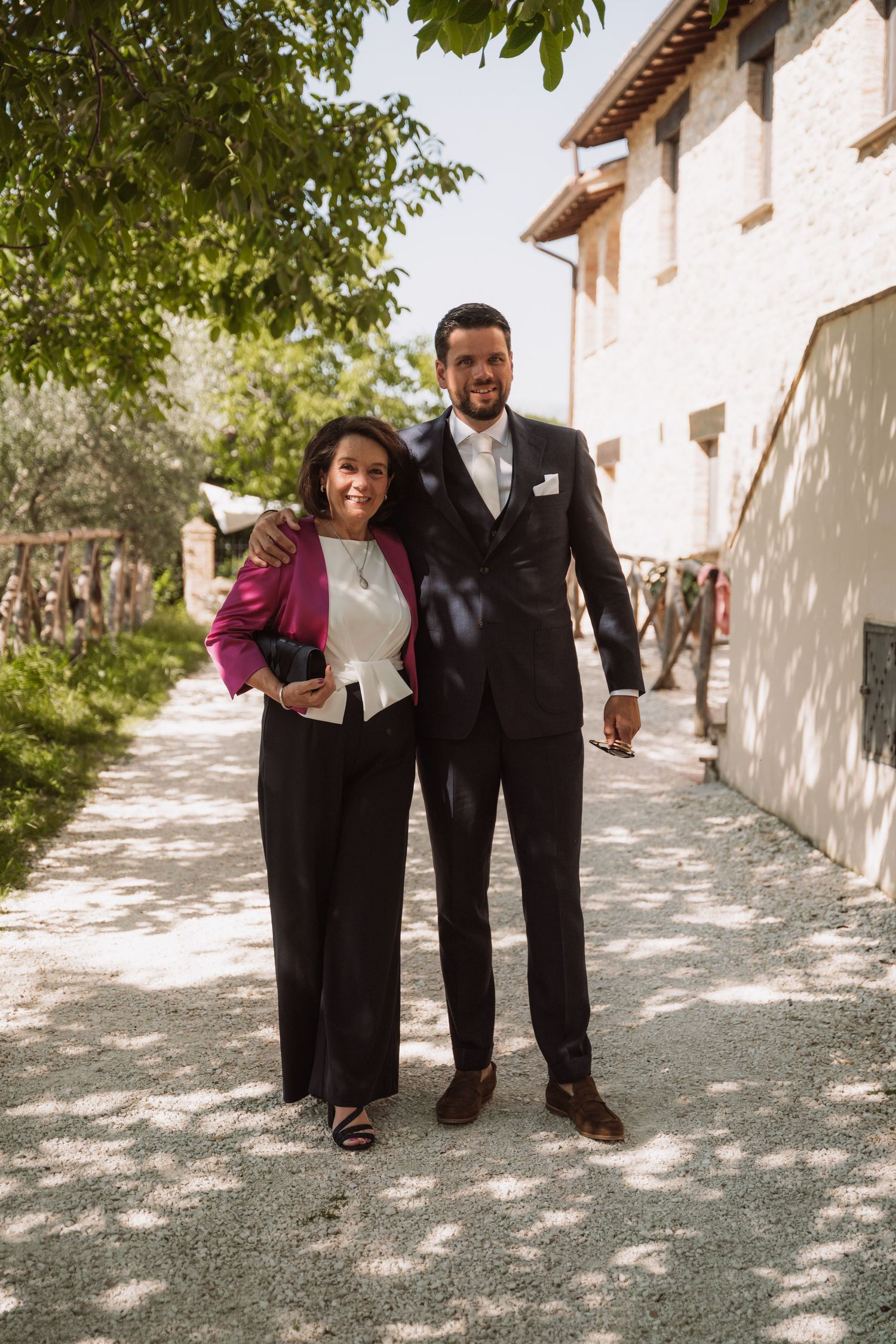 A couple in formal attire walks together along a sunlit path beside a white stucco building on a sunny day.